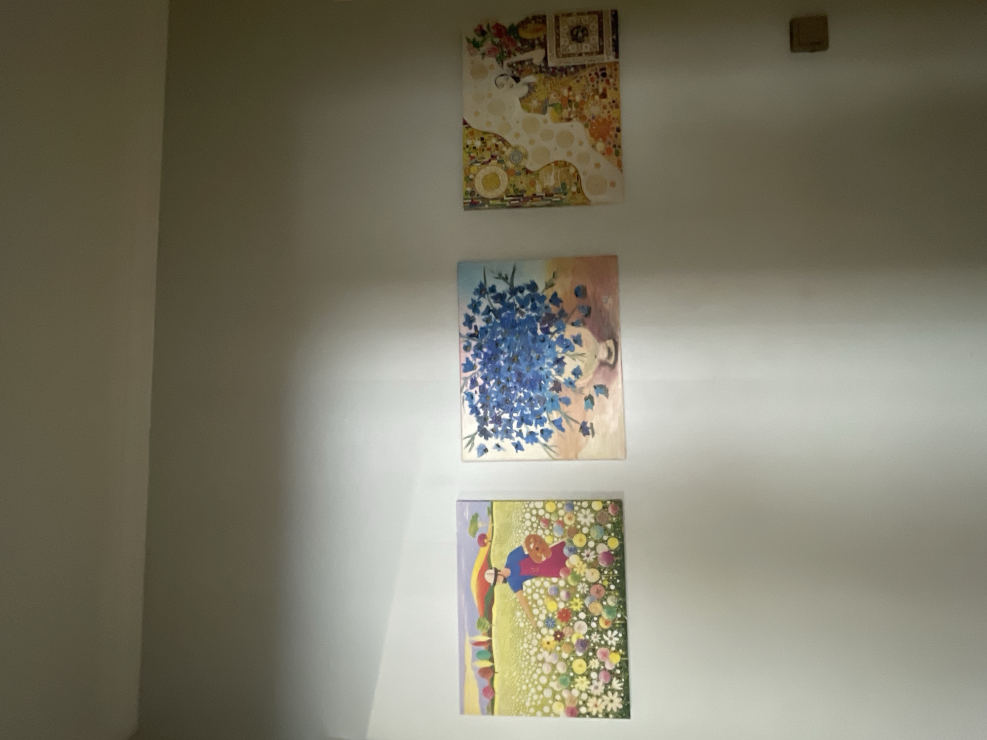 Three framed artworks are displayed on a light grey wall. The leftmost artwork is a landscape painting depicting a person in a blue shirt and red apron, wearing a hat, standing in a field of colorful, circular flowers. The background shows a yellow field, rolling hills with colorful trees, and a purple sky. The middle artwork is a painting of a vase filled with vibrant blue flowers against a light pink and beige background. The rightmost artwork is a square piece with a mosaic-like design featuring abstract shapes, circles, and figures in yellow, orange, and white. There is a light switch on the lower right side of the wall. The scene is indoors, in what appears to be a residential or gallery setting. The location in Almaty, Kazakhstan is not specified by any visual cues in the image.
