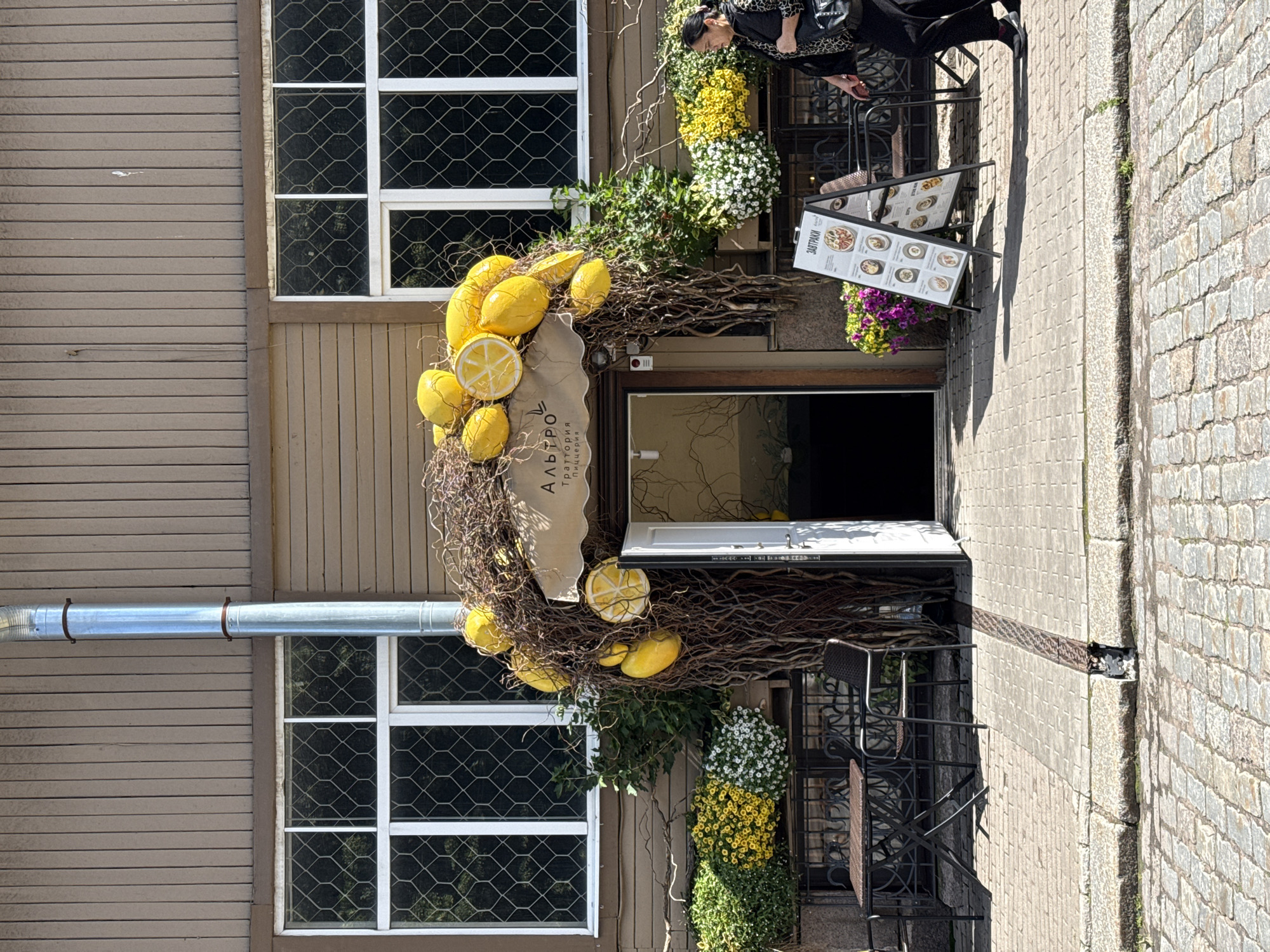 The image shows the exterior of a restaurant with a whimsical lemon-themed entrance. The building is made of wood-paneled siding, and has two large windows with decorative diamond-patterned glass. A prominent silver downspout runs vertically along the side of the building. The entrance is adorned with a large, arching display of branches, intertwined with numerous yellow lemon props and some sliced lemon props. A sign above the entrance reads "Альтро Траттория Пиццерия." To the left of the entrance, there is a table and chair set outside, with planters filled with yellow and white flowers. To the right of the entrance, a woman is walking past, wearing a patterned jacket and carrying a bag. A standing menu board is positioned on the sidewalk in front of the restaurant. The foreground is paved with cobblestones. This scene is located in the city of Vyborg, Russia, likely on one of its historic streets given the architecture and cobblestone paving.