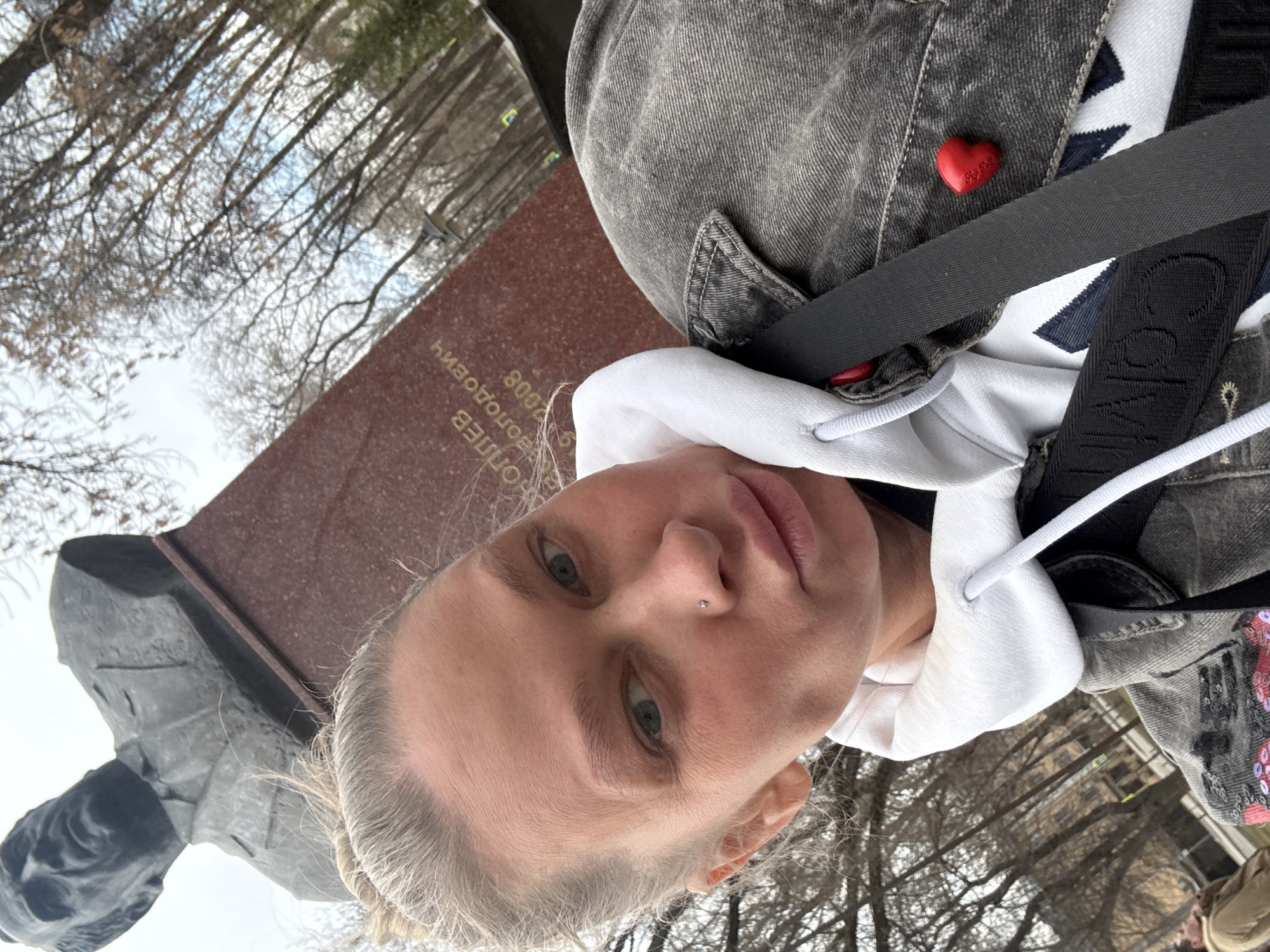 A woman is taking a selfie outdoors, likely in Perm, Russia. She is standing next to a monument, possibly to N.G. Slavianov (based on the inscription that includes his last name and the year 2008). The woman is wearing a gray denim jacket with a red heart pin, a white hoodie, and a black strap with visible white lettering that could be part of a bag or harness. In the background, trees are visible, suggesting a park or wooded area. The image is taken at an angle, with the camera tilted.