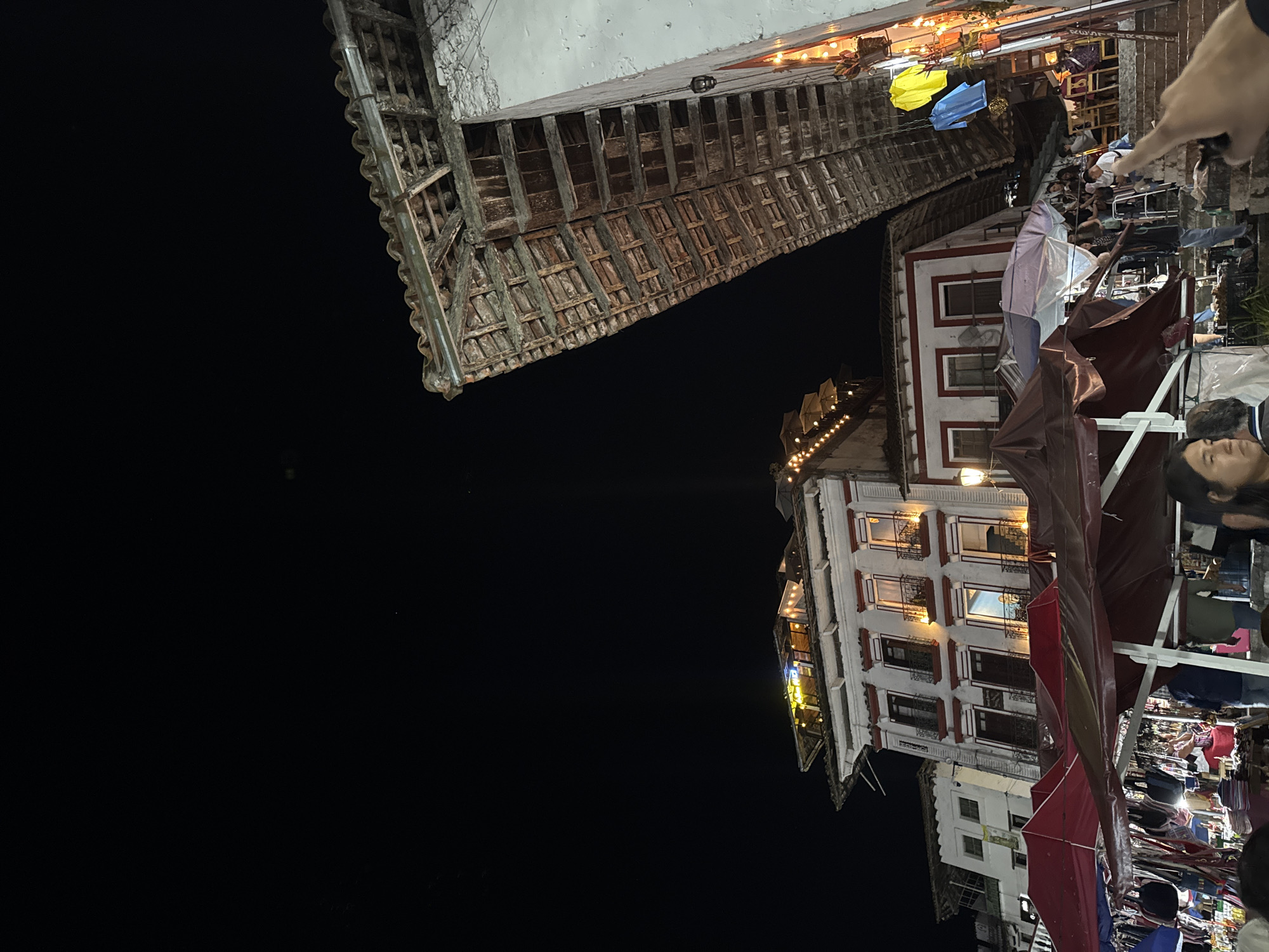 At night in Cuetzalan, Mexico, a photograph captures buildings and market activity. On the right, a multi-story building with white facades and red accents is lit by interior lights, showcasing windows with dark railings. Below, a lively market scene unfolds with people milling around, tents erected, and merchandise displayed under them. The building above has visible aging, with a wooden structure extending overhead. In the upper center, lights hang above, with colorful decorations between the buildings. The sky is dark.