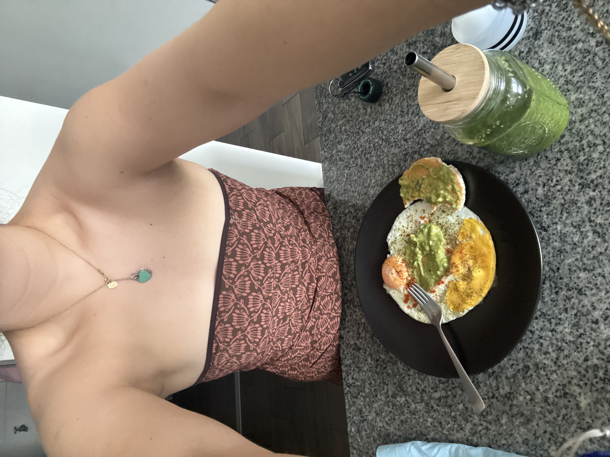An overhead selfie-style shot captures a person and their breakfast in an indoor setting, likely a kitchen or dining area, in Mexico City, Mexico. The person's outstretched arm, shoulder, and upper torso are visible on the left, holding the camera. They wear a brown top or dress with a light-colored, possibly floral or botanical, pattern. A delicate gold chain necklace with a small gold charm and a distinct teal heart-shaped pendant adorns their neck.
On a speckled grey countertop, a black plate holds a meal. It features two fried eggs, one with a visible sunny-side-up yolk and the other primarily egg white, both topped with a generous amount of green avocado or guacamole spread and sprinkled with red paprika and black pepper. A piece of toast or bread, also with the green spread, is on the plate. A silver fork rests beside the food.
To the right of the plate, a clear glass jar with a bamboo lid and a metal straw contains a vibrant green smoothie or juice. A set of keys with a black and green fob lies on the countertop further back. The background shows a white wall or surface and a section of dark flooring. The bright, even lighting suggests it is daytime, indicating a morning or early afternoon meal. No text is visible, and the person's expression is not discernible from the partial view. The scene conveys a calm, domestic moment of preparing to enjoy a healthy meal.