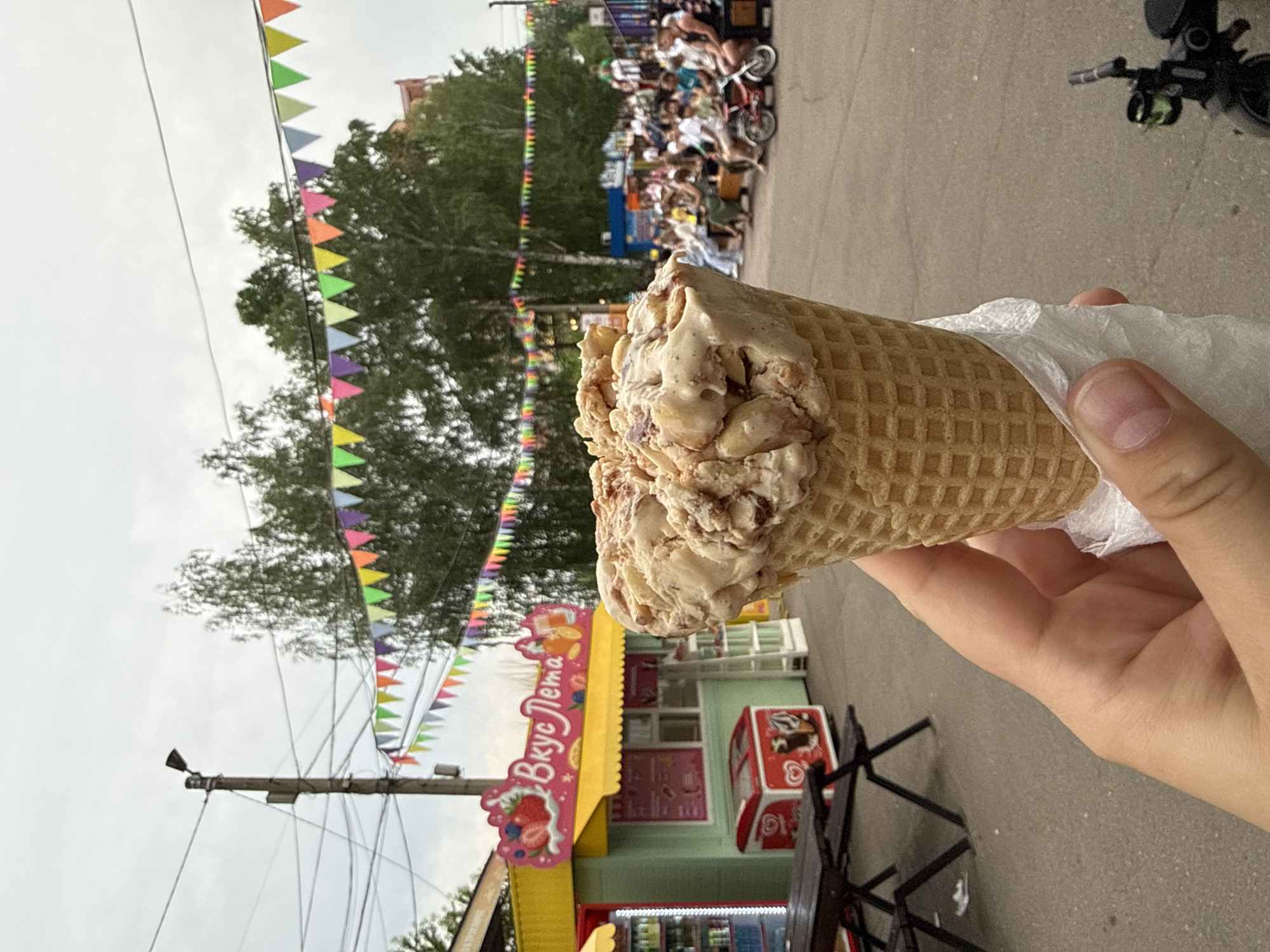 A close-up, first-person perspective shot captures an ice cream cone held in a hand against a backdrop of a fairground or park setting. The ice cream, a generous scoop of a light brown flavor with visible chunks of nuts and caramel, sits in a waffle cone. A white paper wrapper peeks out from under the cone.
In the mid-ground, a small, light green kiosk with a yellow awning is visible, featuring a sign with Russian text that translates to "Summer Taste" and decorated with images of fruits like strawberries and oranges. A red refrigerator with the Coca-Cola logo is placed in front of the kiosk. To the left, a dark wooden picnic table and benches are present.
Further in the background, colorful triangular flags are strung across the scene, creating a festive atmosphere. Trees with lush green foliage dominate the upper mid-ground. A crowd of people is seated on benches and bleachers in the distance, some with children and bicycles, suggesting an outdoor event or gathering. A lamppost with wires is positioned on the left side of the frame. The ground is paved. The overall lighting suggests an overcast day.