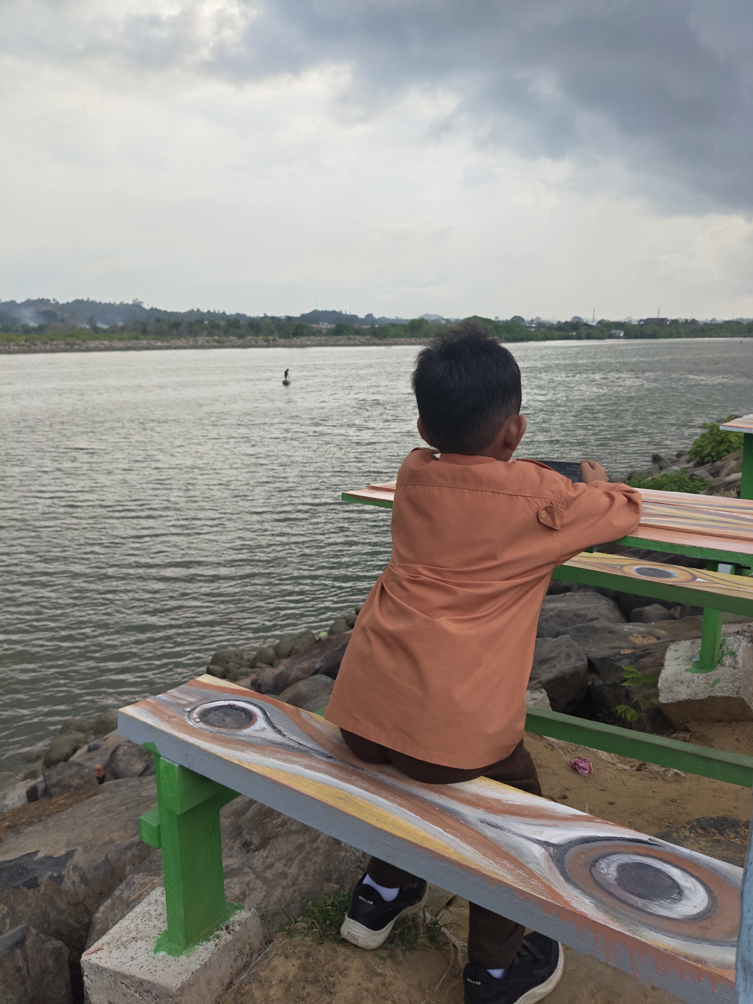 A young boy is sitting on a painted bench overlooking a body of water, likely near Lhokseumawe, Indonesia, based on the location information provided. He is wearing a brown uniform with an orange shirt and black shoes with white socks. He is facing away from the camera, towards the water, and appears to be holding a mobile phone. The bench is painted with a pattern resembling wood grain, and its supports are painted green. Behind the bench are large rocks and sand. In the background, there is a body of water with a person standing on what seems to be a stand-up paddleboard. The sky is overcast.