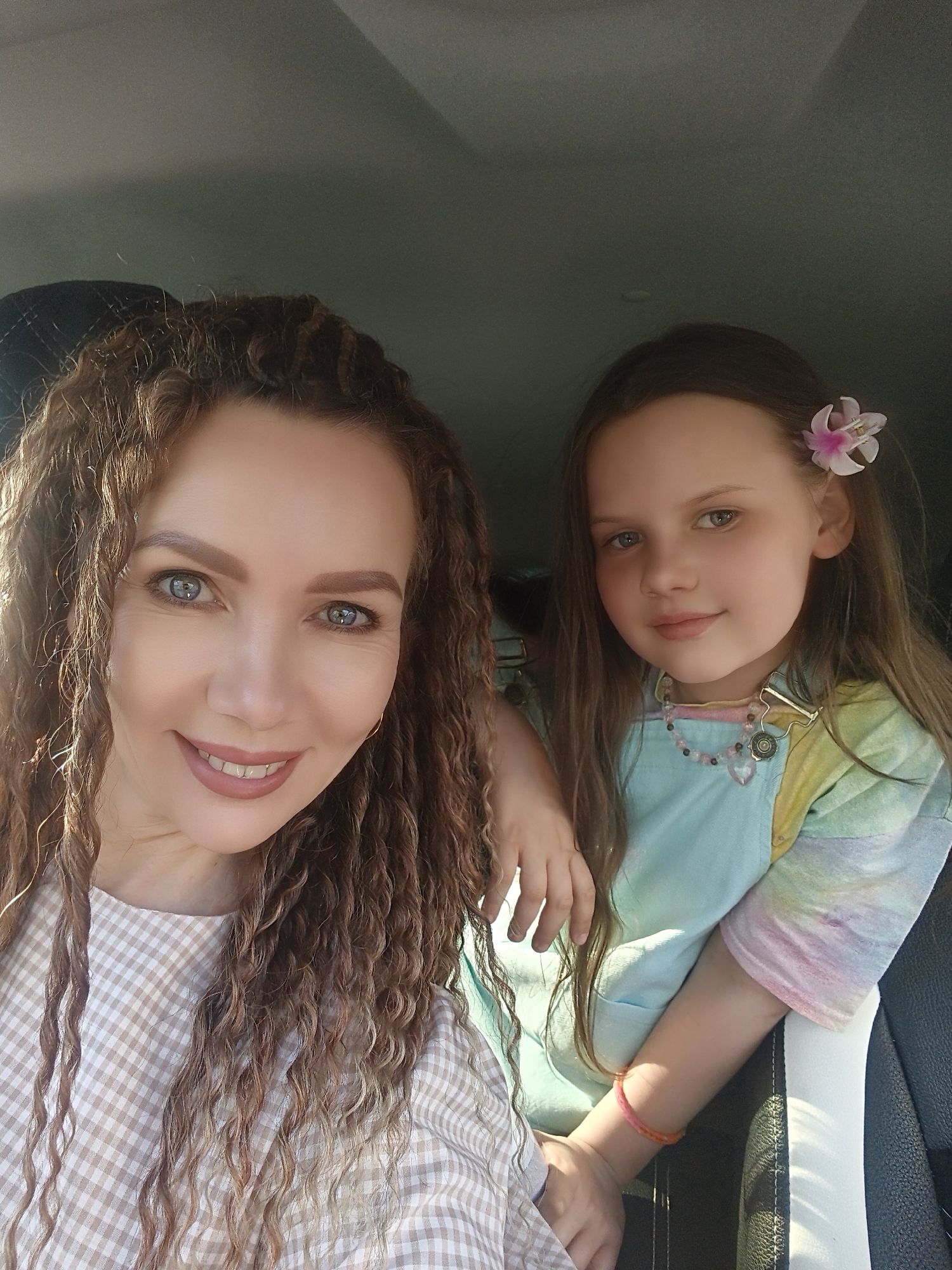 A selfie of a woman and a young girl taken inside a vehicle, likely a car. The woman, on the left side of the frame, has light skin, gray eyes, and curly brown hair. She is wearing a light-colored checkered shirt. The girl, positioned to the right, has light skin, blue eyes, and straight brown hair with a flower pinned in it. She wears a light blue top with tie-dye sleeves and a beaded necklace. Both are looking directly at the camera. The background shows parts of the car's interior, including the headrest of the seat and the ceiling. The location is Unknown, Unknown.