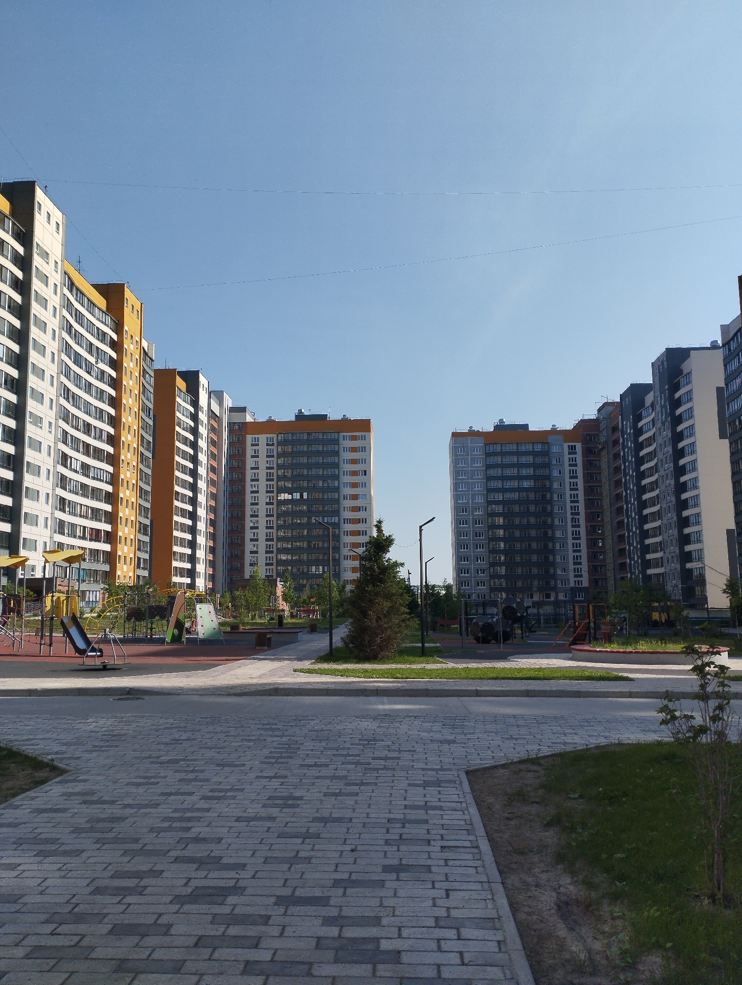 The image depicts a modern residential courtyard in Sadovyy, Russia, on a clear day. Multiple high-rise apartment buildings, characterized by their multi-colored facades of white, yellow, gray, and orange, dominate the background. These buildings feature numerous windows and balconies, indicating a densely populated area. In the foreground, a paved walkway constructed of light gray and dark gray rectangular blocks leads into the scene. To the left, a children's playground is visible with various structures, including slides, swings, and climbing frames, set on a red rubberized surface. Trees and small shrubs are scattered throughout the courtyard, with some grass areas. The sky is a bright, clear blue, suggesting fair weather. No people are present in the image.