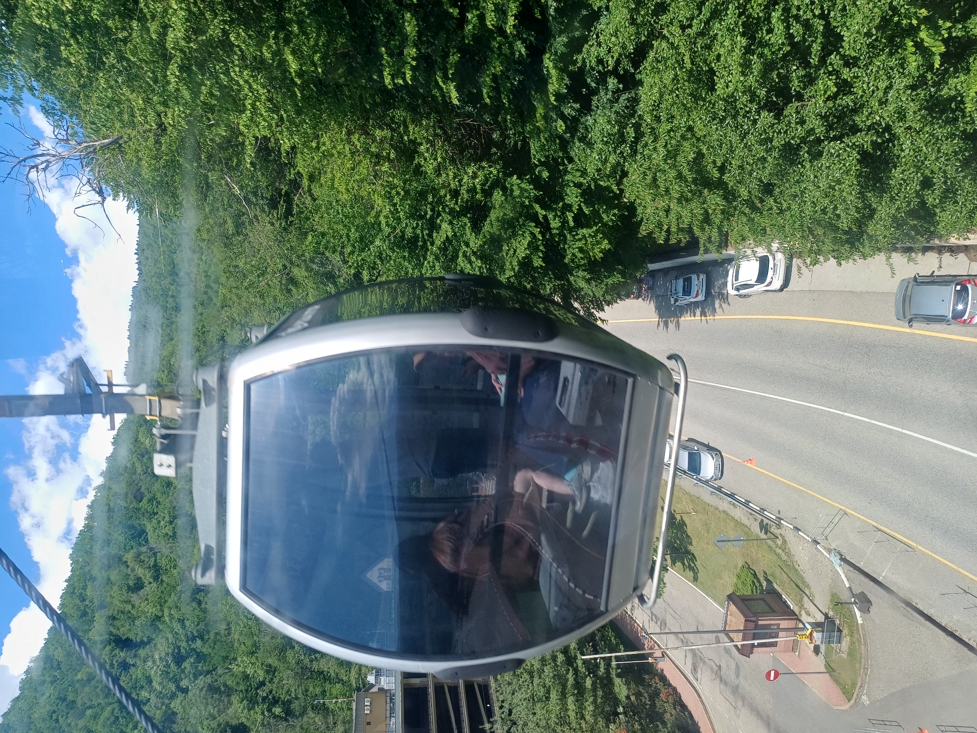 This image presents a high-angle view from inside a gondola lift cabin, rotated counter-clockwise so the horizon is on the left and the ground on the right. The scene depicts a sunny day in Estosadok, Russia, with clear blue skies and fluffy white clouds.
In the foreground, the silver frame and large window of the gondola cabin are prominent. Reflections of people, including one with long hair and another's lower body, are visible on the window, indicating occupants looking out. A thick gondola cable and part of the lift mechanism are visible on the left.
Below, a paved road with yellow and white markings winds through a landscape dominated by lush green trees covering a hillside. Several cars are visible on the road: three light-colored cars are parked along the roadside, and a dark grey/silver car with a roof rack is parked further along. Another silver car is near a small brick building and traffic light structure on the right side of the road.
Notable details include a white triangular road sign with a red border and a "No Entry" symbol near the small building. Another triangular sign, indicating a steep incline/descent, is reflected on the gondola's window. The activity captured is a gondola ride, offering panoramic views of the forested terrain and the road below. The bright daylight suggests it is daytime.