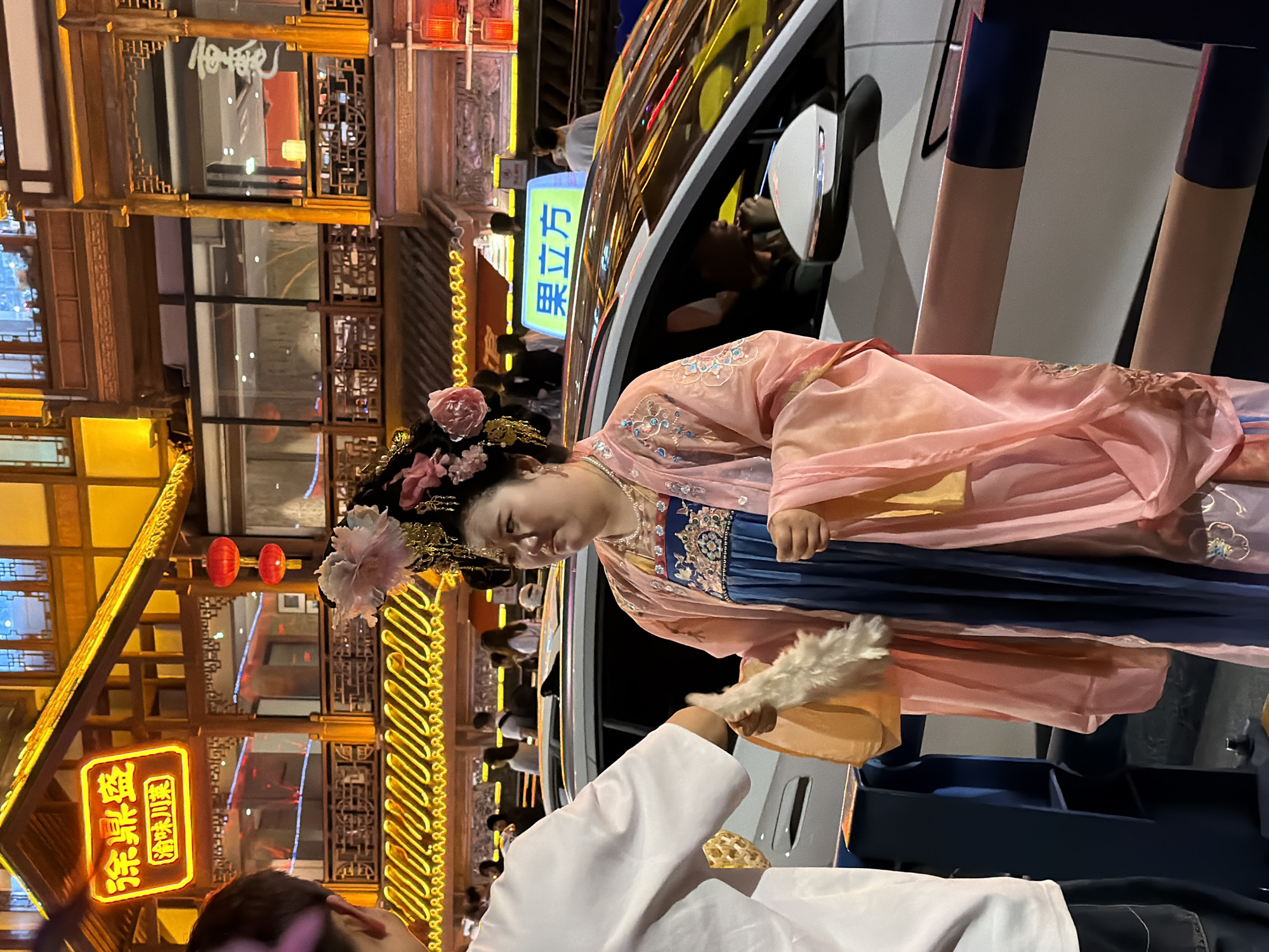 A sideways, medium outdoor shot shows a woman in traditional Chinese dress standing outside a modern car on a city street at night. The woman is in the middle of the frame, facing the lower left corner of the frame, and is slightly blurred. She is wearing a navy-blue dress with gold accents, and a light-pink, sheer outer garment with embroidered flowers on the sleeves. Her dark hair is pulled up and adorned with flowers and gold ornaments. She holds a white fan with feathered trim in her right hand. She is standing near a modern, white car with a black-trimmed side mirror. A person wearing a white jacket is in the lower left corner of the frame, near the woman. The background features a traditional Chinese building with wooden beams and decorative lights. There is a sign with Chinese characters and a brightly lit storefront. The location is Yuzhong District, China.