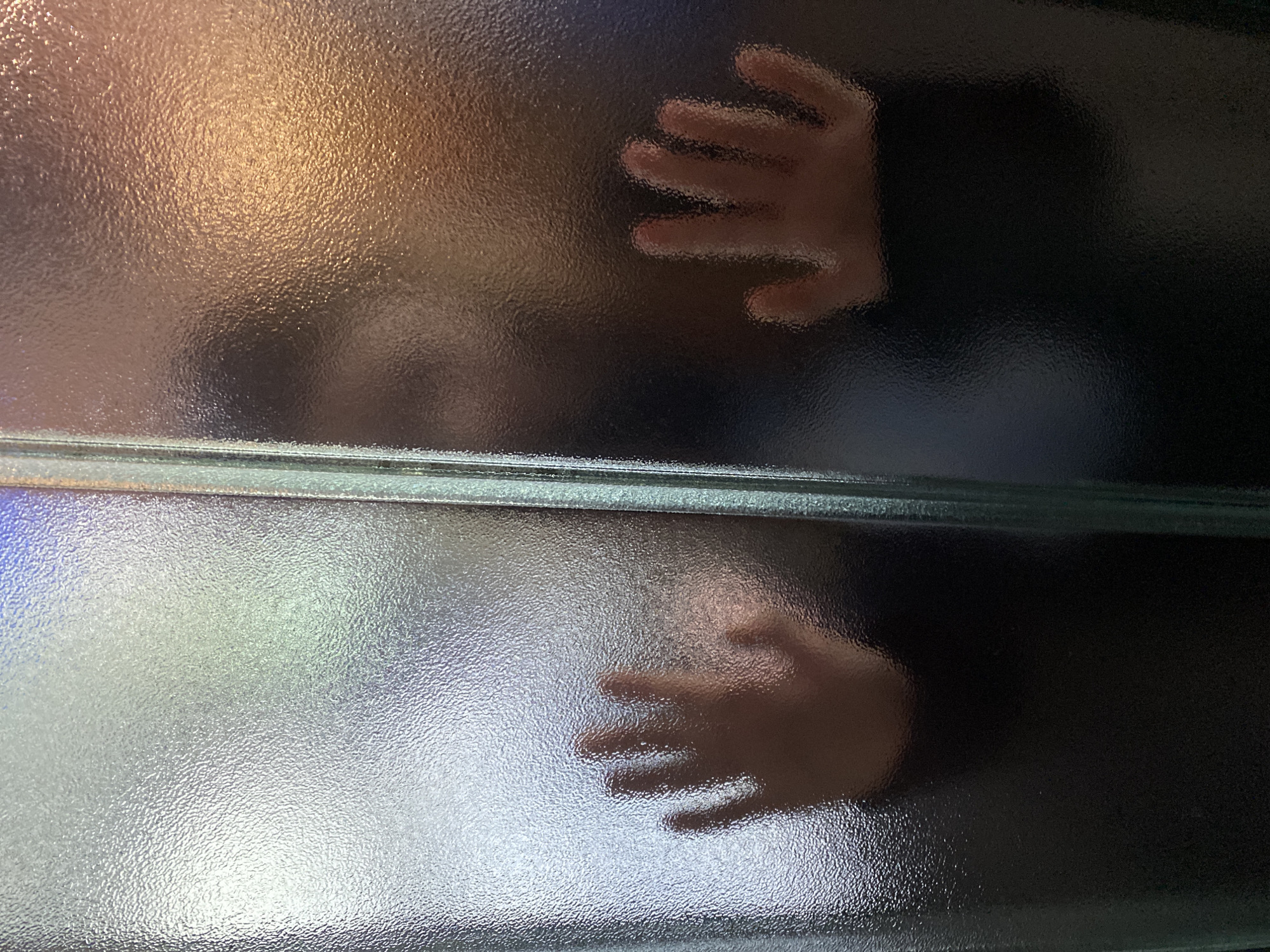 A close-up image shows two hands and a blurred face pressed against a textured glass partition or window. The partition consists of two panes, separated by a horizontal frame. The upper pane has a warm, golden glow, while the lower pane is a cooler silver. The hands are indistinct, suggesting they are behind the glass. The face is also blurred, making it difficult to discern any identifying features. No information indicates the location of the image in Yekaterinburg, Russia. The overall image suggests a sense of obscured presence or a hidden observation point.