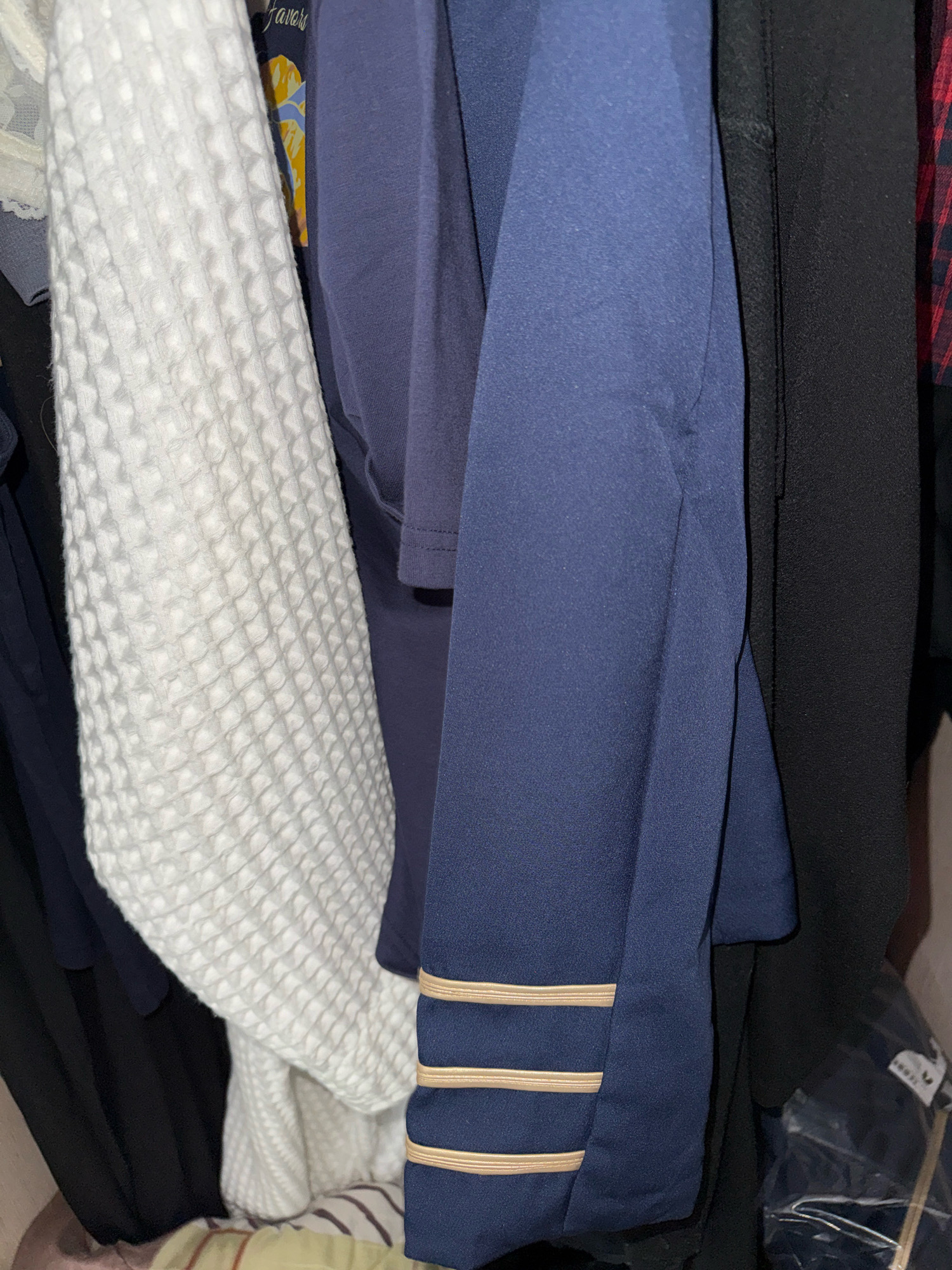 The image shows a close-up view of several items of clothing hanging together. On the left, a white waffle-knit fabric, possibly a robe or towel, is prominent. Next to it, a dark blue garment with yellow lettering, partially obscured, is visible. To the right of that, a solid dark blue sleeve with three horizontal light brown stripes near the cuff is the most detailed item. Behind this, a black garment is visible, and further to the right, a red and black plaid fabric. The clothing appears to be in a closet or wardrobe. The scene is indoors, with no indication of location within Kaliningrad, Russia. There are no people, actions, or interactions depicted.