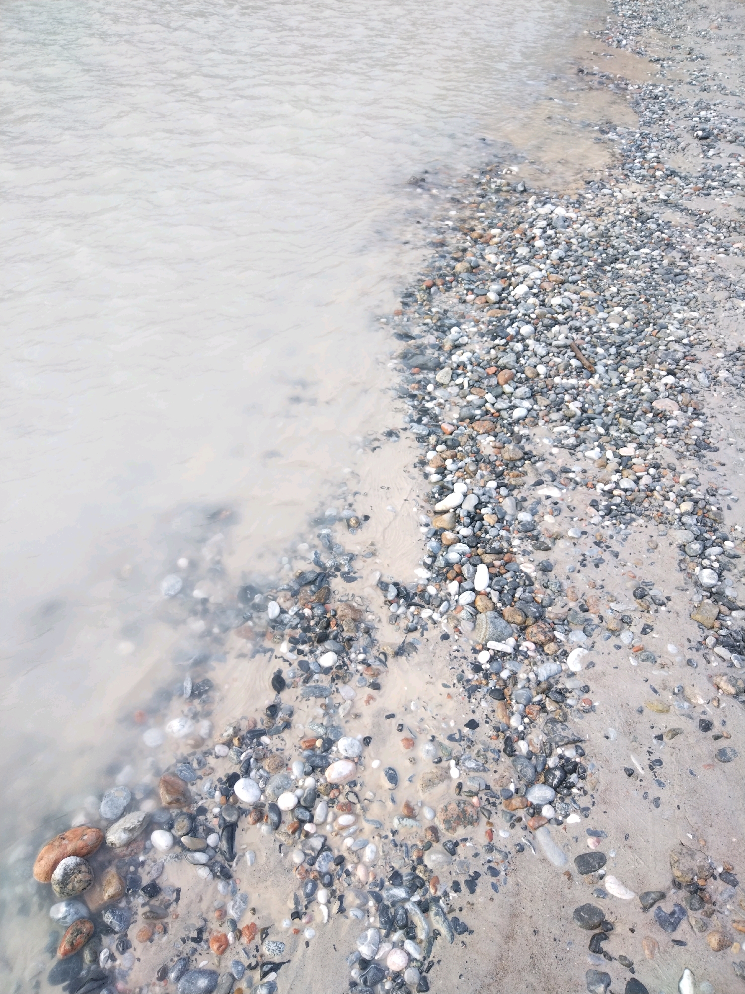 The image depicts a rocky shoreline or riverbank. The foreground is dominated by a mix of sand and numerous smooth, rounded pebbles of various sizes and colors, including grey, white, brown, and some reddish-orange hues. The pebbles are wet, indicating proximity to water. The left side of the image shows a body of murky, light-brown water with subtle ripples. The water's edge is indistinct, blending into the sandy and pebbly terrain. The background is a continuation of the same pebbly and sandy shore, with the water extending further. There are no people or specific structures visible in the image. The overall environment suggests a natural water body's edge, possibly a lake, river, or sea. The location in Yordon, Uzbekistan, is not visually evident from the provided image itself, as there are no identifying landmarks or specific geographical features that would confirm this location.