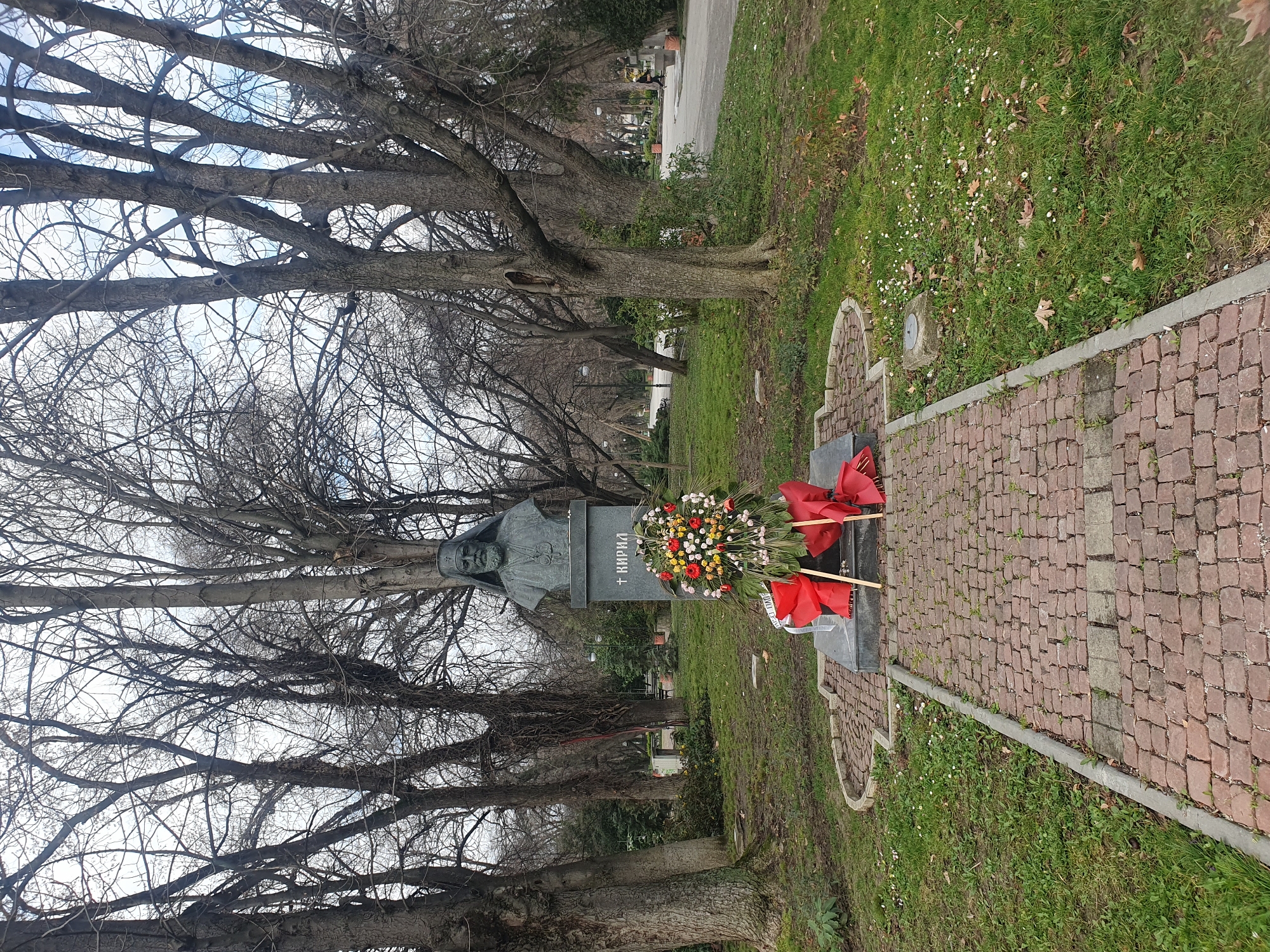 The image presents a bronze bust of Patriarch Kiril of Bulgaria atop a rectangular grey stone pedestal, situated within the City Garden (Tsar Simeon Garden) in Plovdiv, Bulgaria. The bust depicts a mature, bearded male figure wearing a clerical kamilavka and a panagia. The pedestal bears the Cyrillic inscription "+ КИРИЛ". Directly in front of the pedestal, a large, circular floral wreath, primarily composed of red, yellow, and white flowers with prominent red ribbon streamers, rests on a dark grey slab. Additional loose red ribbons are visible on the ground immediately below the wreath.
A reddish-brown cobblestone pathway approaches the monument, bordered by a low concrete curb. Surrounding the path and monument is green grass, sparsely covered with fallen brown leaves and small white flowering plants. The scene is dominated by numerous tall deciduous trees with bare branches, indicating a late autumn or winter season, set against a light grey, overcast sky. In the mid-ground to the right, a paved park pathway extends into the distance, with a single individual in dark attire walking away from the camera. Distant streetlights and a park bench are visible further along this path. The ground is largely clear of debris, with minor irregularities in the grass.