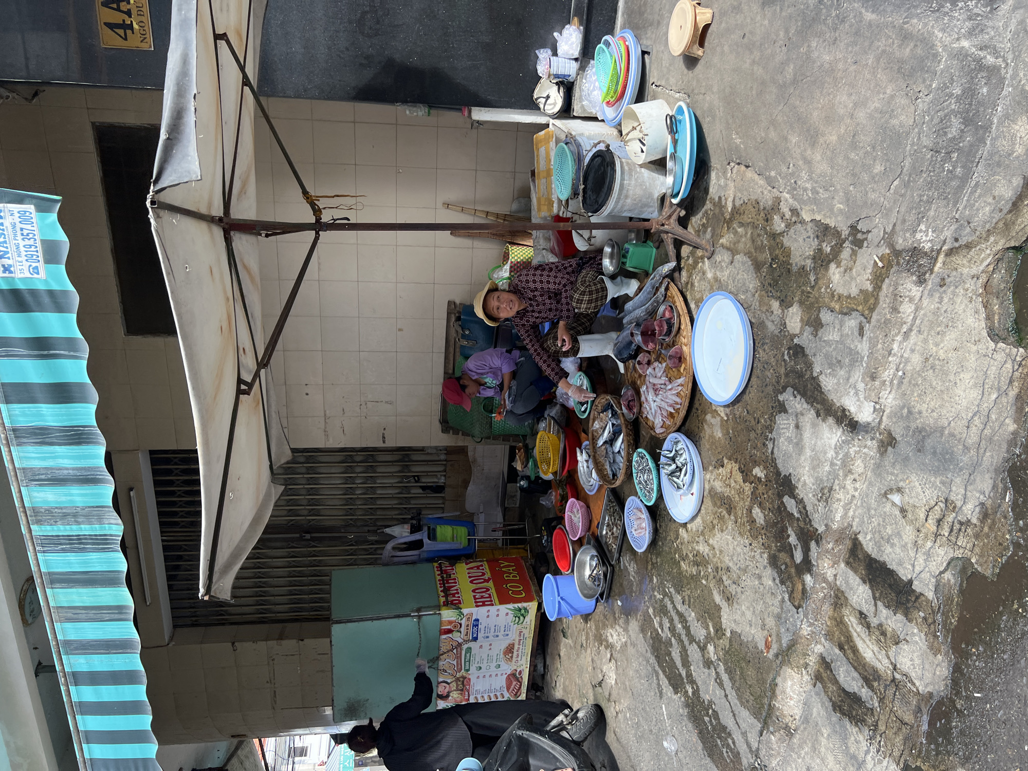 The image depicts a street market scene in Nha Trang, Vietnam. The setting appears to be an outdoor market stall situated on a wet, cracked concrete surface, likely a sidewalk or alley. The stall is covered by a large, off-white umbrella supported by a metal pole.
In the foreground, numerous types of fish are laid out on various plates, baskets, and trays for sale. Some fish are whole, while others are cut into pieces. There are also cutting boards with raw fish on them. Several plastic bowls and buckets are also present, some containing water or fish.
Two women are present. One woman, wearing a black outfit and her hair tied up, stands behind a green refrigerated display case. She appears to be a vendor. The other woman, wearing a red cap and purple shirt, is seated on the ground, possibly assisting or another vendor. A third woman, wearing a straw hat and a patterned shirt and pants, is seated on the ground in front of the fish display. She is also a vendor and is looking towards the camera.
The background shows the facade of a building with tiled walls and a metal roll-up door. An awning with teal and black stripes is visible above the stall. A sign with Vietnamese text and a phone number is also present. A motorcycle is parked to the left of the scene. The overall impression is a bustling, open-air fish market where vendors are preparing and selling their fresh catch.