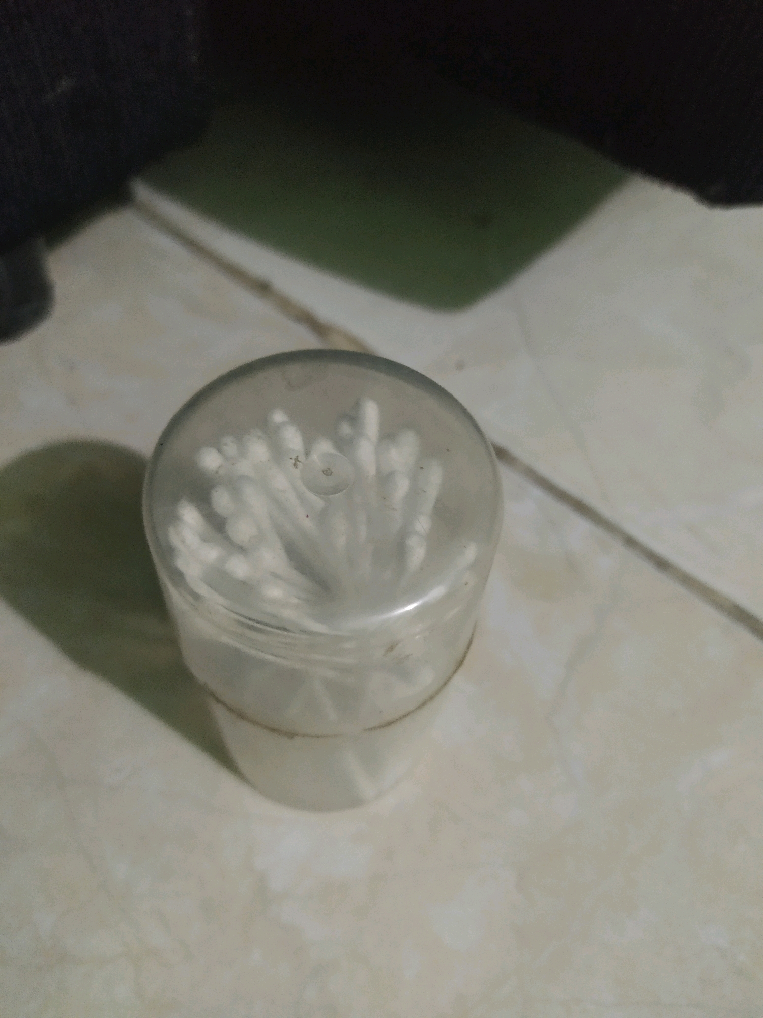 A close-up, high-angle shot shows a clear plastic container filled with white cotton swabs. The container is cylindrical with a rounded lid. The cotton swabs are neatly packed inside, with their white cotton ends pointing upwards. The container is resting on a tiled surface that appears to be a floor. The tiles are light-colored with visible grout lines. In the background, a dark, out-of-focus object is visible, possibly clothing or furniture. A shadow is cast on the floor to the left of the container, suggesting an overhead light source. The image is not explicitly set in Sroyo, Indonesia; this detail is a user-provided context.