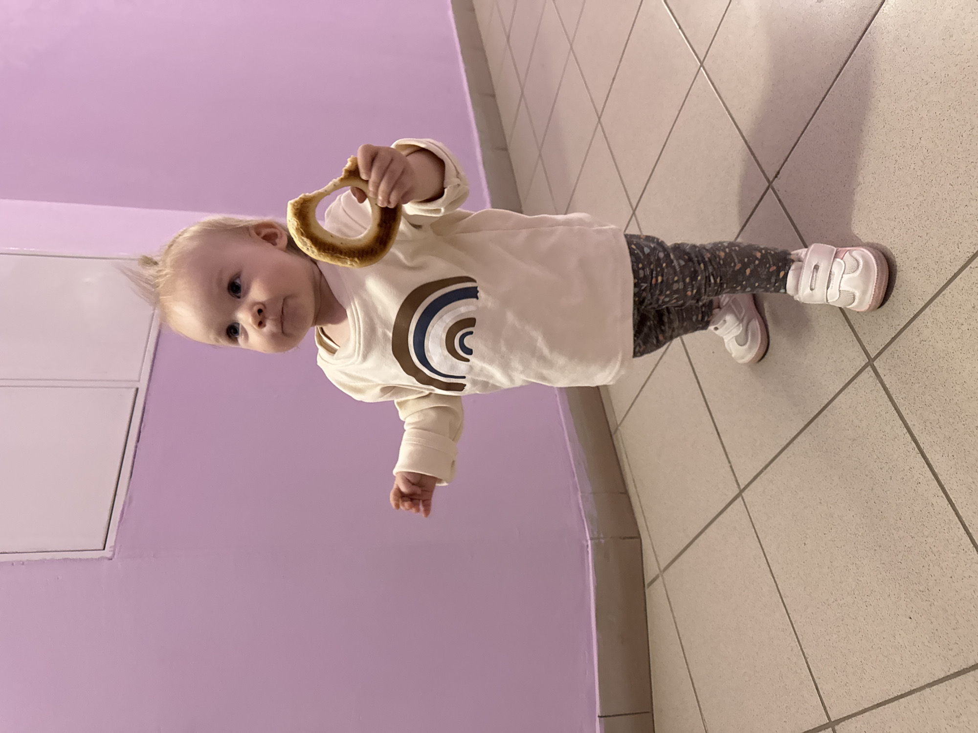 A young child stands in a hallway in Chaikovsky, Russia. The hallway has a tiled floor and lilac-colored walls. The child holds a bagel in their right hand, while their left hand hangs at their side. They wear a cream-colored shirt with a rainbow design, leggings, and white shoes. A white panel is visible on the left wall.
