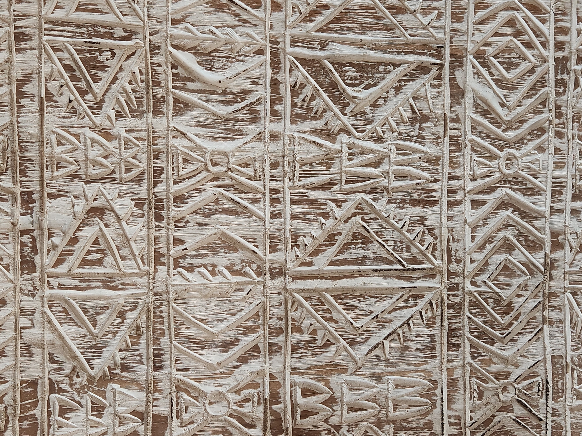 The image shows a close-up of a textured, carved wooden surface with a white, distressed finish. The carving is in horizontal bands, with geometric patterns and stylized motifs that appear somewhat tribal or folkloric. The wood grain is visible beneath the white paint, giving the surface a rustic and aged appearance. There are no people, animals, or any other discernible objects or activities present in the image. The context of Maitland, United States of America, is not visually represented in the photograph itself.