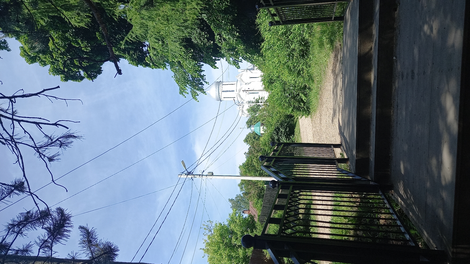The image captures an outdoor scene in Zvenigorod, Russia, showing an approach to the Assumption Cathedral on Gorodok. In the immediate foreground, dark metal railings flank a set of dark wooden steps, partially shaded, leading onto a light-colored dirt path. Dark pine branches are visible in the upper left, and bare deciduous branches in the upper right, against a light blue sky. The path is bordered by dense green bushes and trees. On the left, a utility pole with a street lamp and multiple black electrical wires extends upwards. Behind the foliage on the left, a segment of a wooden fence and a building with a red roof are partially discernible. A smaller structure with a green conical dome is situated amidst the greenery to the left of the path, preceding the main edifice. In the central background, the Assumption Cathedral on Gorodok, a white Orthodox church with a prominent silver central dome and smaller silver domes, rises above the surrounding green trees. The sky above is clear with scattered white clouds. No human or animal subjects are present in the scene.