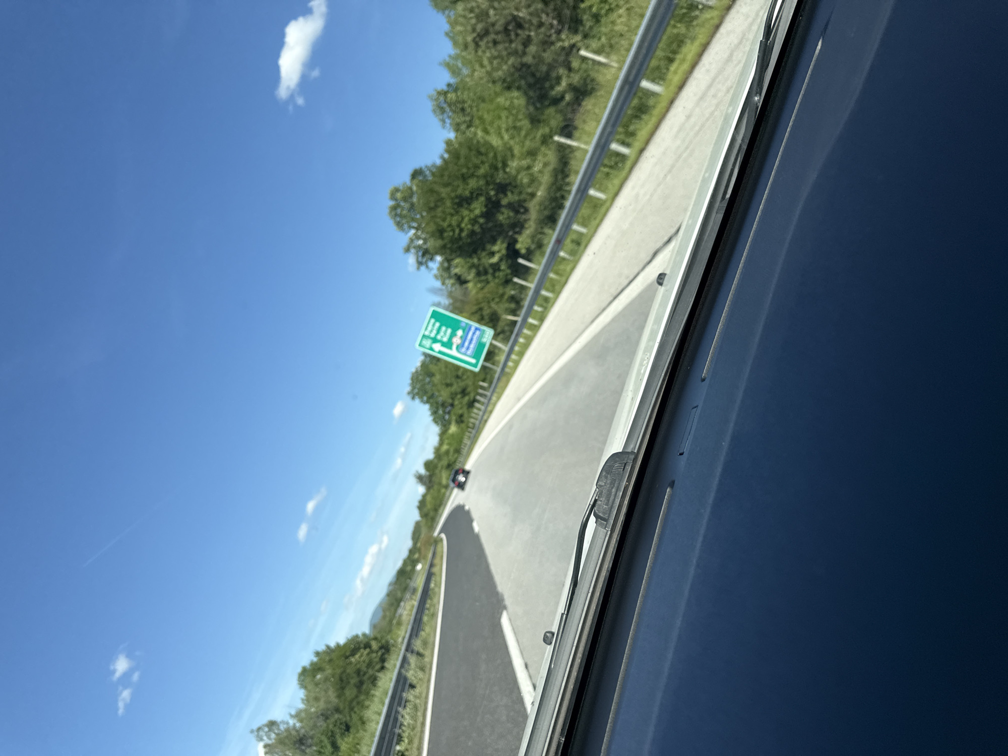 The image shows a view from inside a vehicle driving on a highway. The vehicle is on the right lane of a two-lane highway. There is a car ahead in the same lane. Green foliage is visible on the sides of the highway. A sign indicating directions is visible to the left of the highway. The sky is blue with some clouds. The location is Osikovska Lakavitsa, Bulgaria.