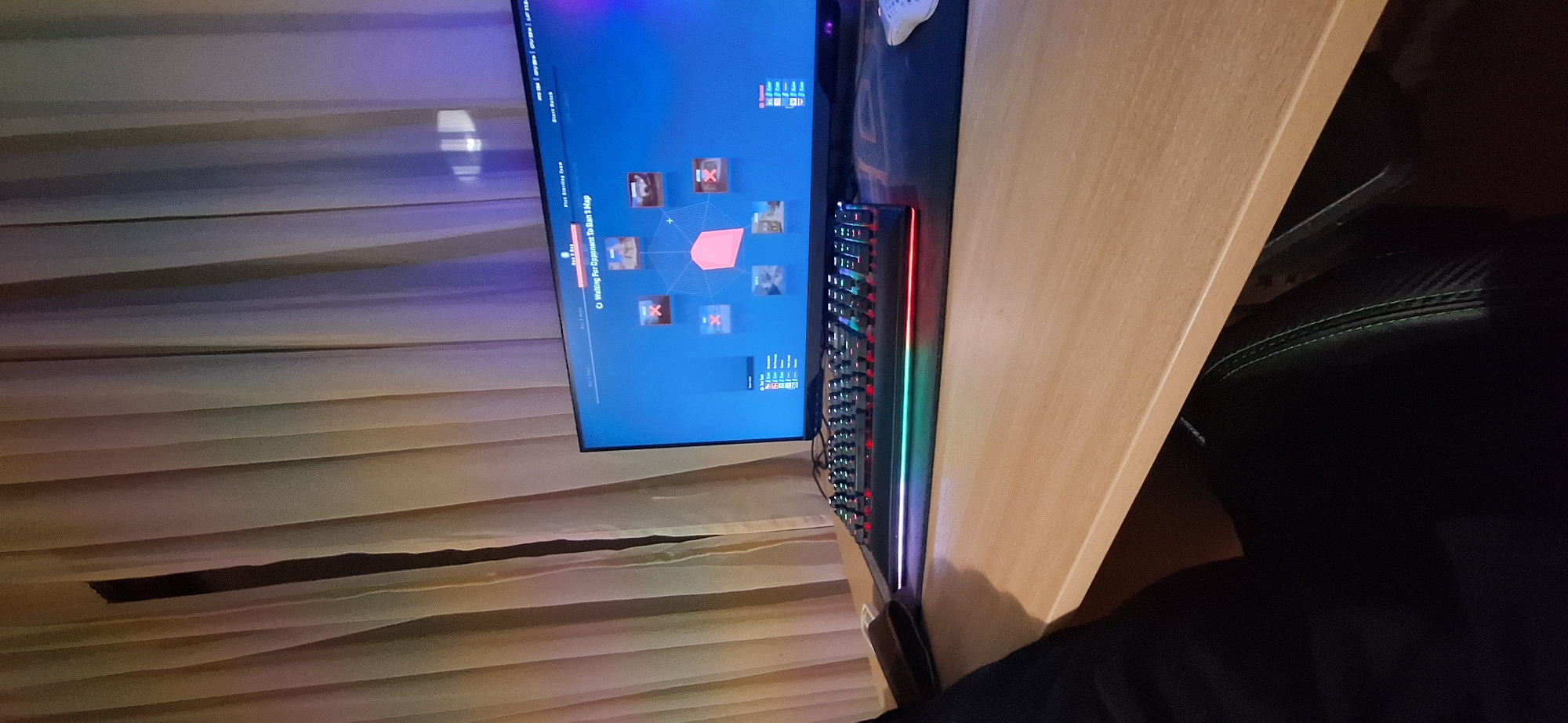 The image shows a gaming setup. A monitor displays a game interface. A keyboard with RGB lighting sits on a light-colored desk. Adjacent to the desk, part of a black chair is visible, with a person wearing black pants sitting in it. A window with tan-colored curtains is in the background. The image does not provide enough information to determine if the location is in Burgas, Bulgaria.