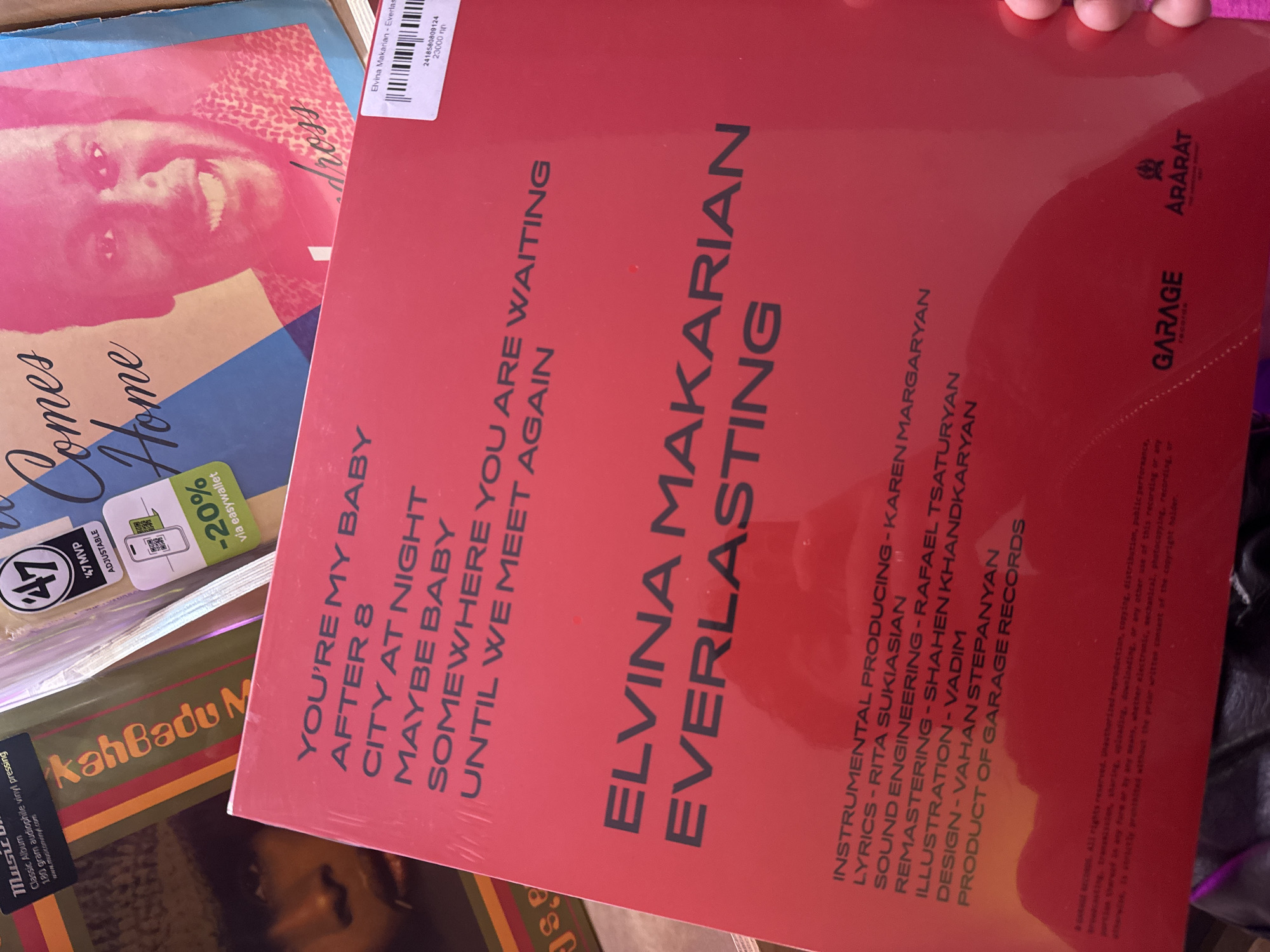 The image shows several vinyl record album covers. The prominent one in the center is red and titled "Elvina Makarian Everlasting," likely the name of the artist and album title. To its left are other albums, including one with the visible titles "Comes Home" and "kah Badu M". Text on the record cover mentions "GARAGE records" and "ARARAT", which suggests a link to Yerevan, Armenia, since Mount Ararat is a significant symbol there. The album's text also includes "INSTRUMENTAL PRODUCING - KAREN MARGARYAN," "LYRICS-RITA SUKIASIAN," "ILLUSTRATION-VADIM," "SOUND ENGINEERING - RAFAEL TSATURYAN," "REMASTERING - SHAHEN KHANDKARYAN," and "DESIGN-VAHAN STEPANYAN." The album cover also has a barcode with the number "2418580809124" and the price "23000 np." The other album is partially visible, and there's a sticker on the packaging mentioning a "20% discount via easywallet".