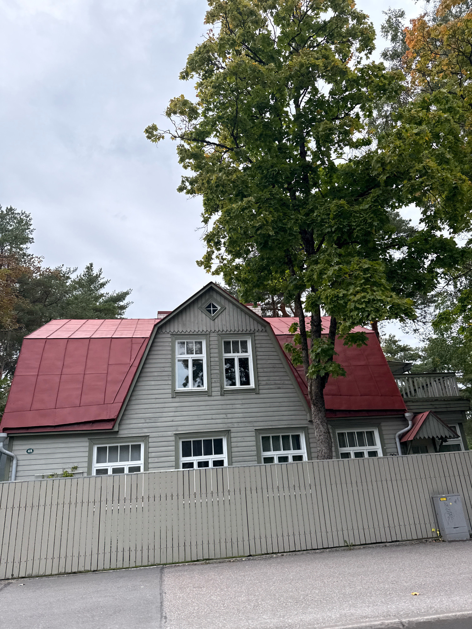 The image shows a two-story wooden house with a red metal roof, located behind a pale grey wooden fence. The house has multiple windows with white frames. A large tree with green leaves partially obscures the right side of the house. The house is painted a light grey color. The sky is overcast. In the background, more trees are visible. A utility box is situated to the right of the fence. A street is visible in the foreground. The house number "48" is visible on the left side of the house. This scene is consistent with a residential property in Tallinn, Estonia, likely in an older neighborhood given the architectural style of the house.