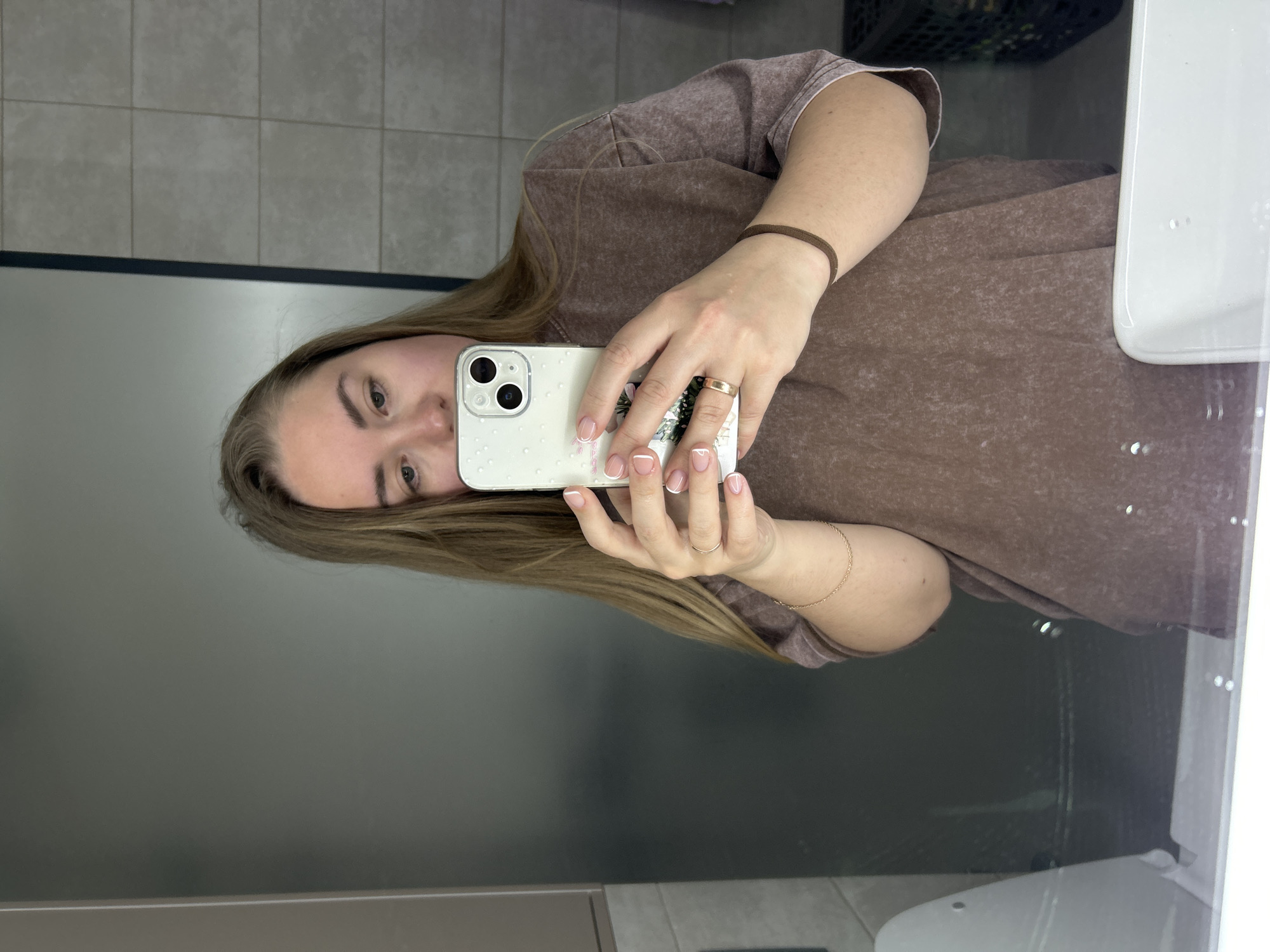 A woman is taking a selfie in a restroom. She has blonde hair and is wearing a brown t-shirt, a brown wristband, and a gold bracelet. Her phone has a white case with a floral design and water droplets on it. Her nails are manicured with a French tip. The restroom has gray tile walls and a white sink and toilet. The location is Chaikovsky, Russia.