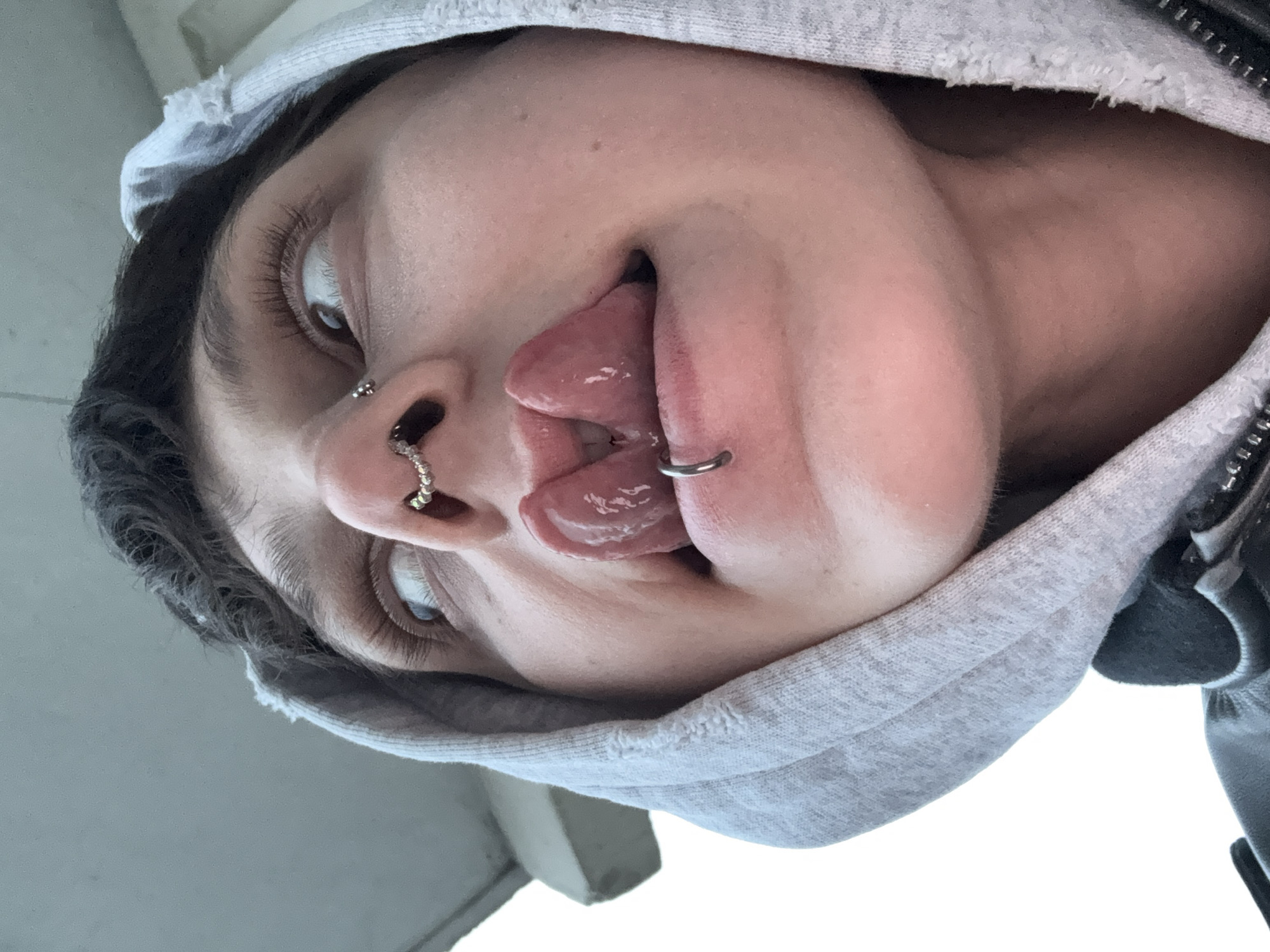 The image shows a person wearing a gray hoodie and multiple piercings. There are piercings in the nose and a ring through the tongue. The background is out of focus and does not provide any evidence to suggest the location is Moscow, Russia.