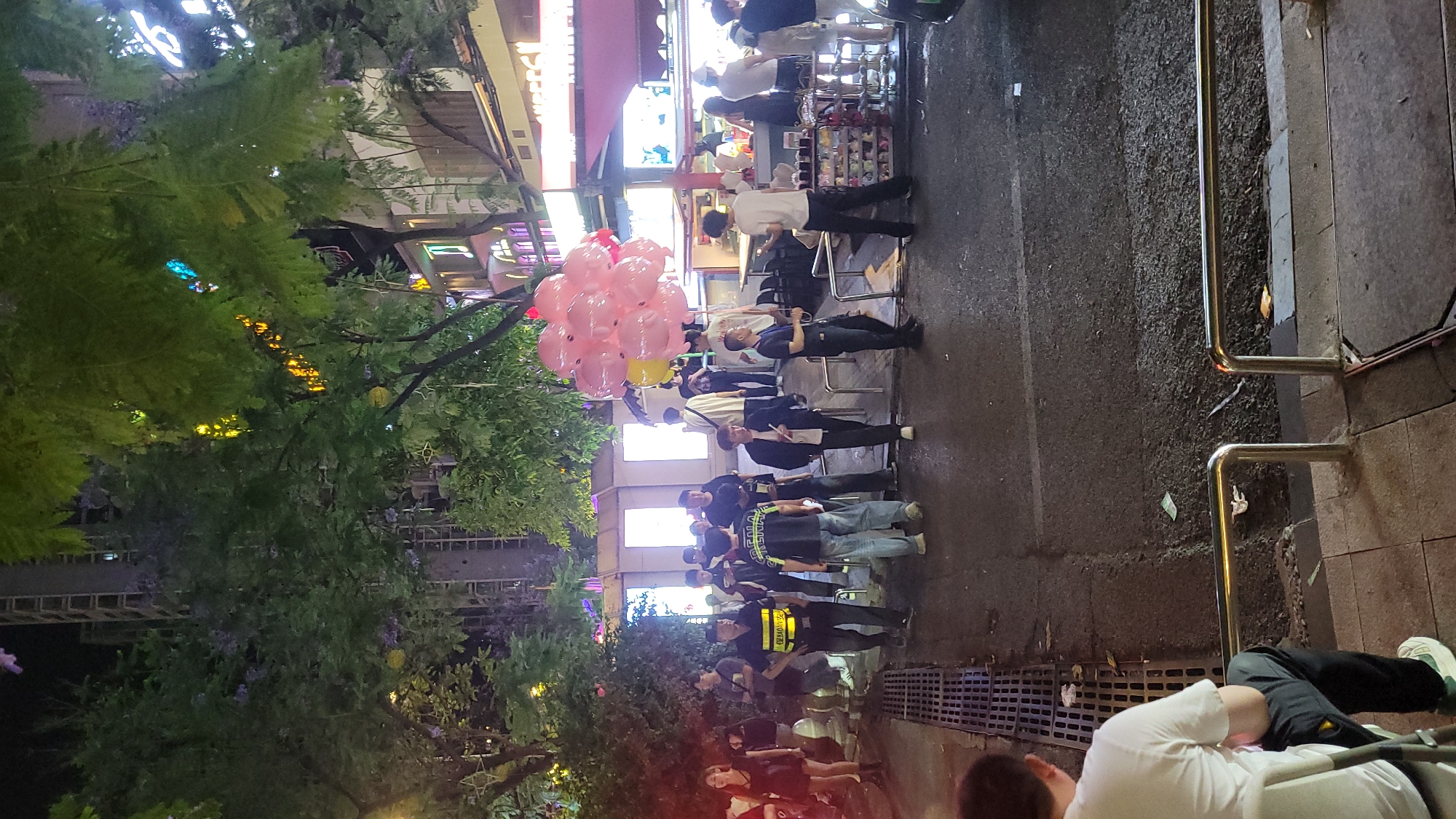 The scene shows an outdoor street scene at night in what appears to be a commercial area in Unknown, Unknown. A group of people is standing on the pavement; many of them are wearing face masks. Some of the people are standing, while others are seated on chairs. A vendor is selling a bouquet of pink balloons. There are trees and vegetation on the left side of the image, with decorative lights in the trees. On the right side, there is a metal railing, and a person is sitting on the ground next to it. The street pavement is wet, and there is a drainage grate visible in the foreground. Several buildings are visible in the background with storefronts and lighting.