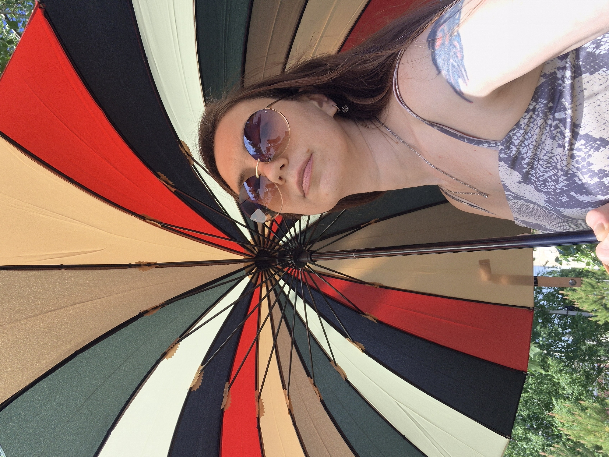 A photograph depicts a woman with light skin, wearing sunglasses and a light-colored patterned tank top, partially visible under a large, multicolored parasol. The parasol is composed of alternating panels in shades of red, black, tan, white, and green. The woman has long brown hair and a tattoo visible on her upper arm. The setting appears to be outdoors, with trees visible in the background, possibly in the city of Chaikovsky, Russia. The woman is holding the parasol above her head.