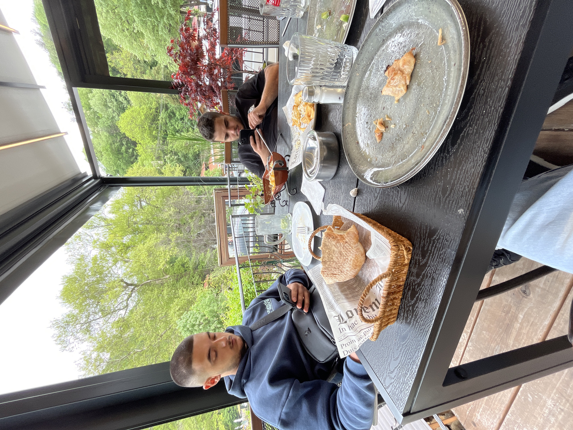 The image depicts a table scene with two men at a restaurant, possibly in Bachkovo, Bulgaria, given the lush green vegetation visible in the background and the architectural style of the surrounding buildings. The table is set with remnants of a meal, including plates with food scraps, glasses, and condiments. One man is looking at his phone while the other is reading a newspaper. A basket with bread is placed on the table, as well as a black leather bag. The restaurant appears to have an open-air terrace or patio with a view of trees and buildings. The lighting suggests it is daytime.