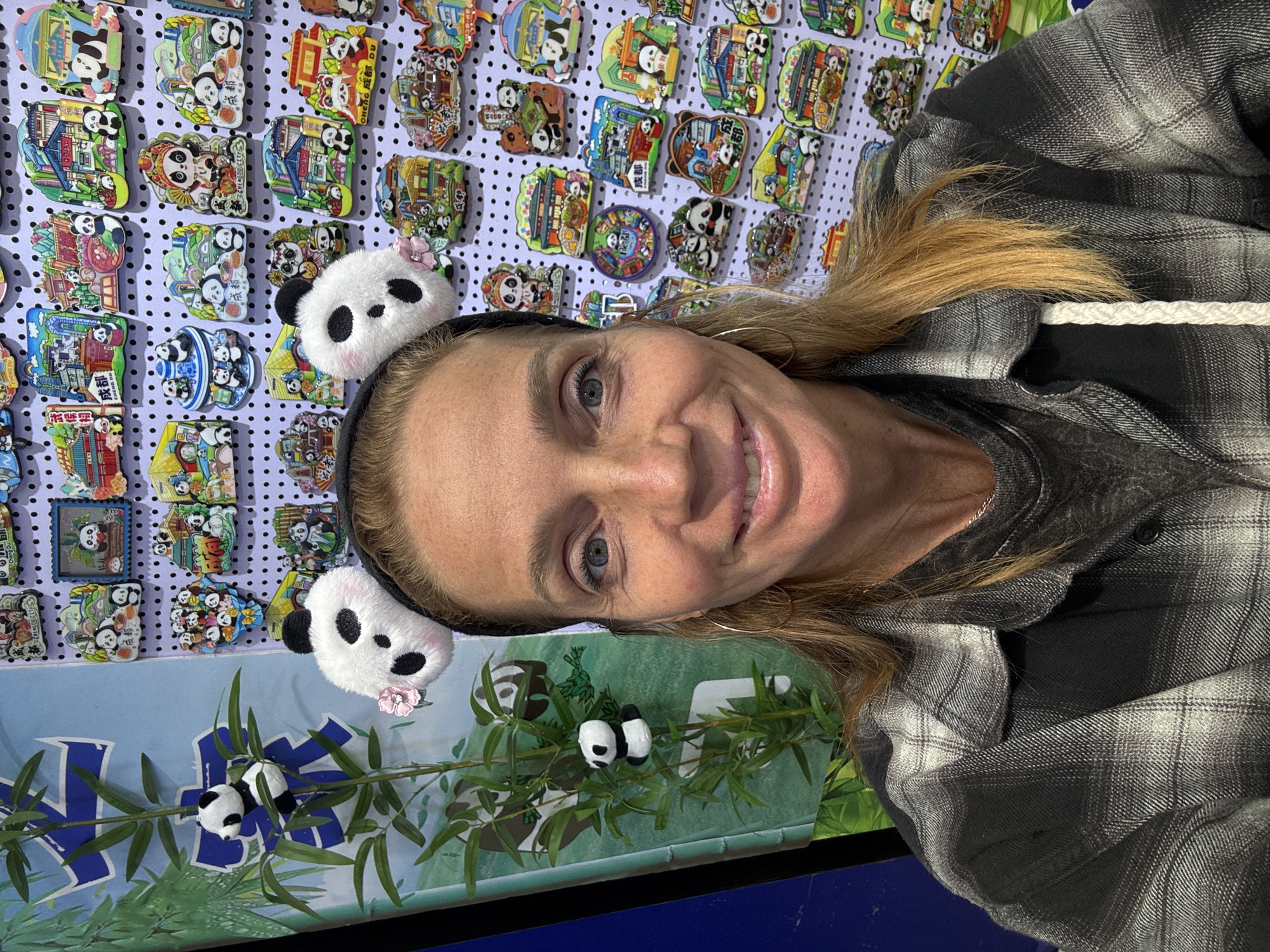 The image shows a selfie of a woman with a panda-themed headband, standing in front of a display of panda-themed merchandise. The woman has blonde hair, blue eyes, and is wearing a gray plaid shirt. The headband has two plush panda heads with pink flowers. Behind her, there is a display board covered with various panda-themed pins and magnets, featuring cartoon pandas in scenes related to Chengdu. Below the display board, there is a backdrop depicting bamboo and small panda figures. The location can be inferred to be related to pandas and Chengdu, likely the city of Jinjiang District, China, based on the details depicted in the merchandise and backdrop.