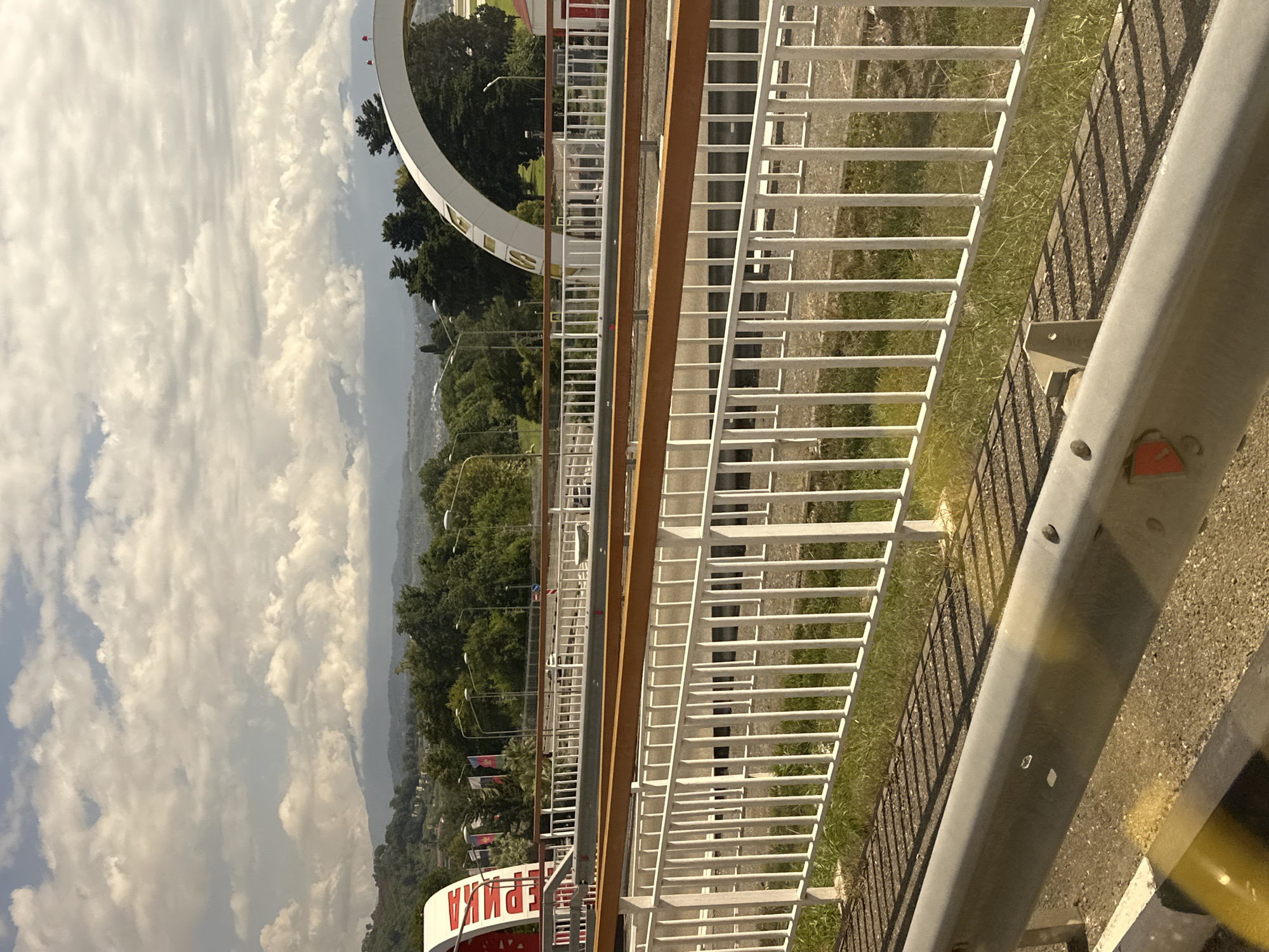 The image depicts a view from a road overlooking a bridge structure in Sochi, Russia. In the foreground, a white metal railing with a wooden top rail is prominent, casting shadows from its vertical bars onto the grassy ground below. Beyond the railing, a highway with traffic can be seen, with cars moving in the distance. Two large, white archways with reddish-colored architectural elements are visible on either side of the road. The left archway has Cyrillic lettering, which appears to read "РОССИЯ" (Russia). The right archway is adorned with small red flags at its apex. Behind the bridge, a lush green landscape with trees and distant mountains under a cloudy sky completes the scene. The lighting suggests it is daytime, possibly late afternoon given the long shadows. There are no people visible in the image.