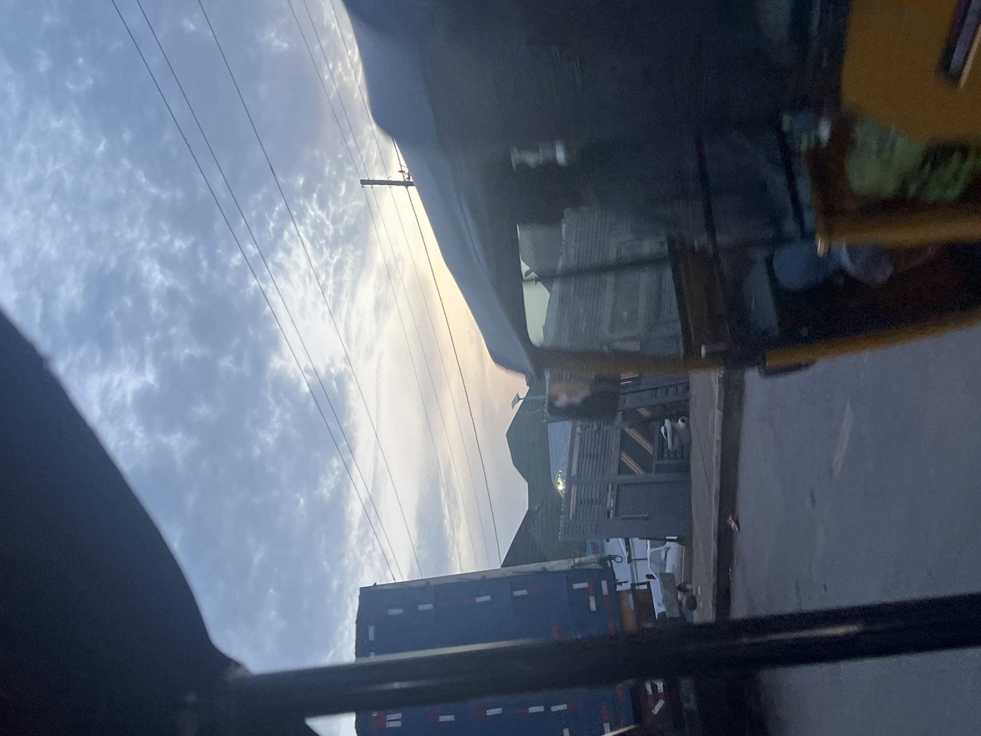 The image depicts a street scene in Jalingo, Nigeria, possibly during dawn or dusk, as indicated by the soft, diffused light and the overcast sky with scattered clouds. In the foreground, a portion of a yellow auto-rickshaw, commonly known as an "okada" or "keke napep," is visible, suggesting the perspective is from within one. The rickshaw's window is partially visible, showing a blurred interior and the dark exterior frame.
To the left, a large blue truck with white and red markings is parked or in transit. Its trailer is tall and rectangular, suggesting it might be carrying goods. Behind the truck, several structures are visible. One appears to be a building with a gray, patterned facade, and another is a grey wall with horizontal slats. Further in the background, rolling hills or low mountains form the horizon under the cloudy sky. Several power lines are strung across the upper part of the frame, supported by poles.
On the street, a white utility vehicle is parked near the grey building. Scattered on the ground near the curb are a few small objects, including what appears to be a red piece of litter. The overall impression is that of an urban or semi-urban environment in Nigeria, captured from a moving vehicle, likely an auto-rickshaw.