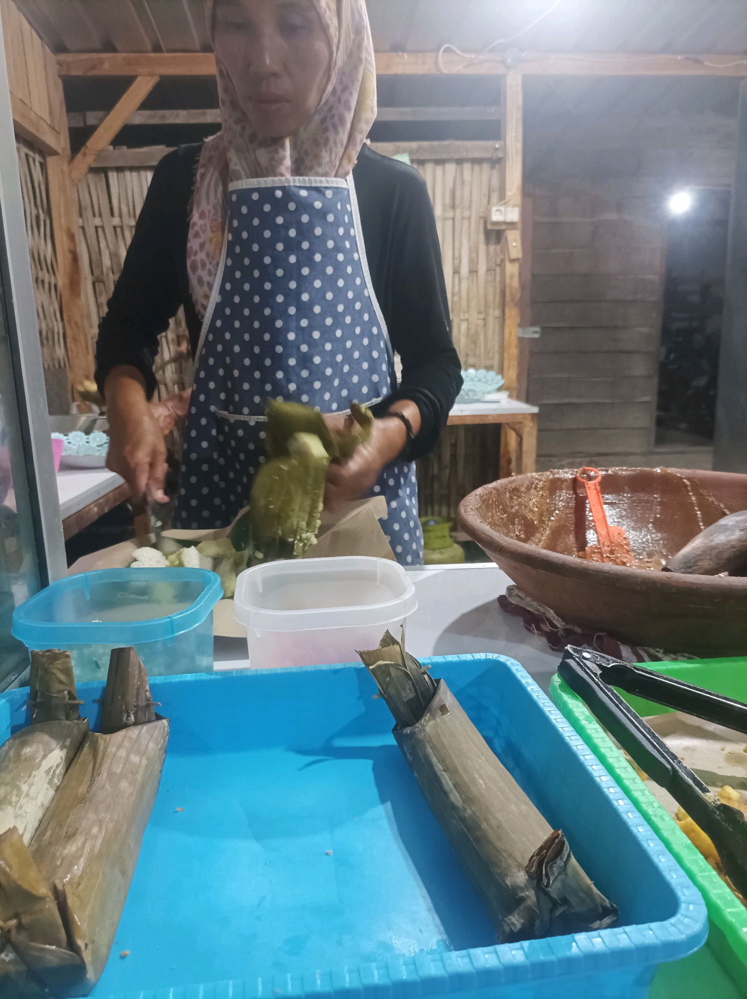 In Sumuragung, Indonesia, a woman wearing a hijab and a blue and white polka-dot apron is seen preparing food. She is in what appears to be an outdoor or semi-outdoor food stall. In front of her, on a white countertop, are two plastic containers, one clear blue and the other clear white, filled with food ingredients. Her hands are actively engaged in wrapping food items in banana leaves. To the right, a large, rustic clay bowl holds a brown, semi-liquid substance, possibly a sauce or condiment, with an orange utensil and a pestle inside. Further to the right, a blue plastic tray holds two food items wrapped in banana leaves, and a green plastic tray contains tongs and more food, partially obscured. The background shows a rustic structure made of wood and bamboo, with lighting from an overhead bulb. The overall scene suggests a culinary preparation in a traditional Indonesian setting.