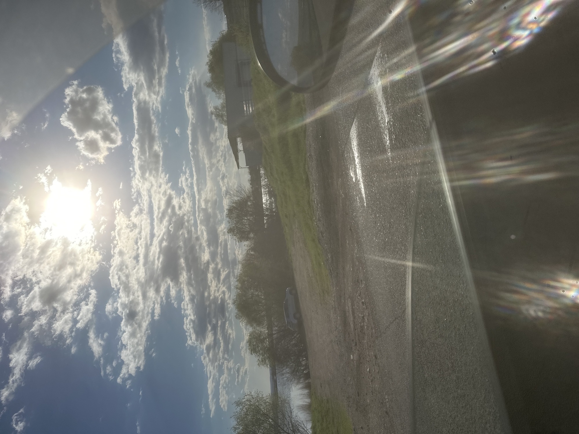 The image appears to be taken from inside a vehicle, possibly a car, looking out through the windshield. The view encompasses a scene with a bright sky filled with scattered clouds and the sun shining through them. Below the sky, there's a strip of land with trees and vegetation. The land appears to border a body of water, possibly a lake or river. A building is visible in the distance, possibly a house or structure on the shore. A side mirror of the vehicle is visible. The reflection of the sky, clouds, and sunlight is visible in the windshield. The location of Burgas, Bulgaria, is not confirmed by the image.