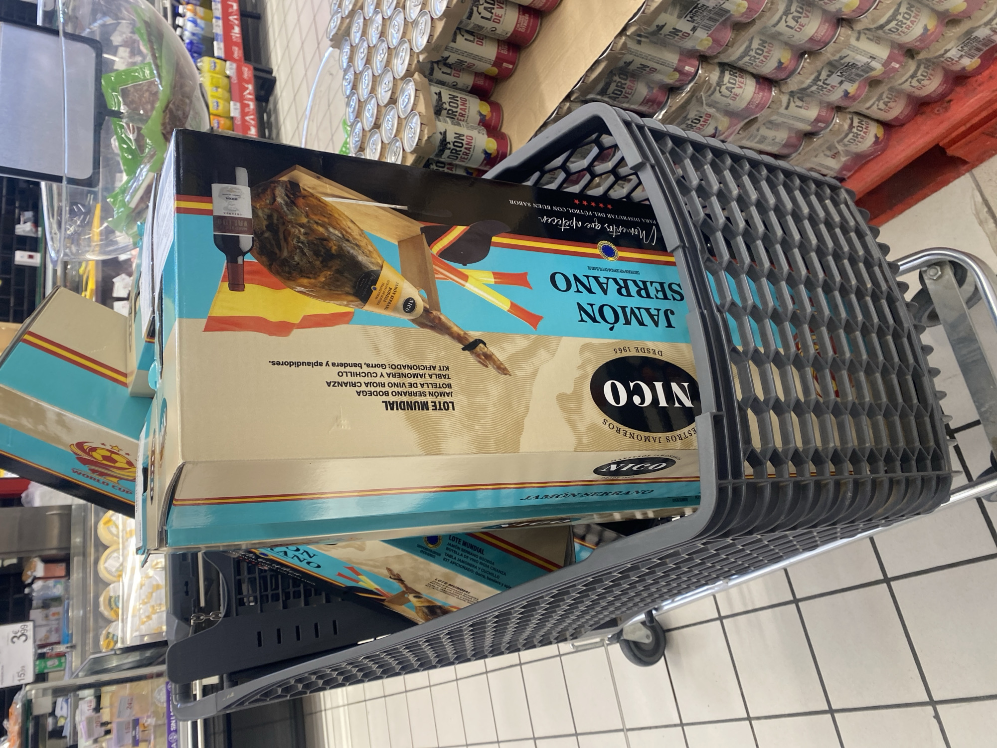 The image depicts a shopping cart filled with several large boxes of "Jamón Serrano" (cured ham). The boxes are decorated with Spanish flags and images of ham. In the background, shelves of canned drinks are visible, along with some supermarket displays. The flooring is tiled. The shopping cart is positioned in an aisle of a grocery store or supermarket. The overall scene suggests a retail environment in Spain.