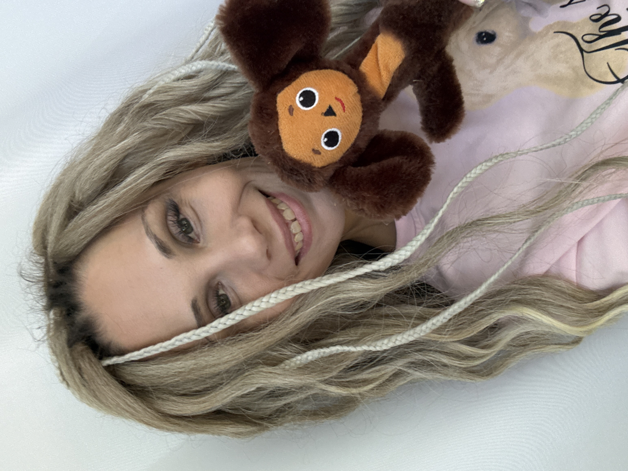 A woman with long, blonde wavy hair with white braids is lying down and smiling at the camera. She is holding a brown and orange stuffed animal that resembles a cartoon character, possibly Cheburashka. She is wearing a light pink top with writing on it. The background appears to be a plain, light-colored surface. There is no information to indicate that this image was taken in the city of Zavolzhye, Russia.