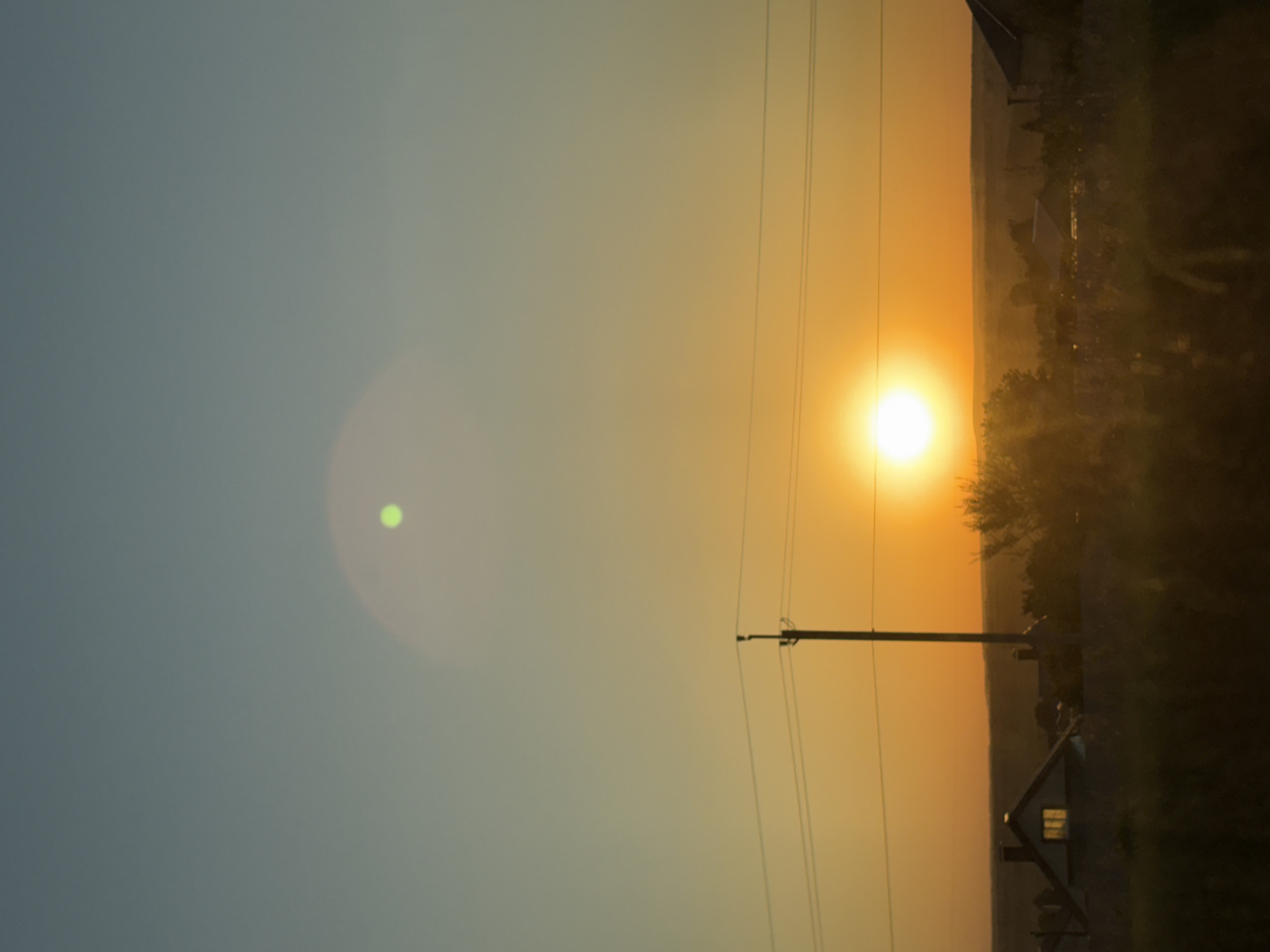 Captured from Bol'shiye Klyuchishchi, Russia, the landscape unfolds under a vibrant sunset. The sun, a luminous orange orb, dominates the sky, partially obscured by a cluster of trees and silhouetted buildings on the horizon. Overhead power lines cut across the sky, intersecting with a utility pole in the foreground. In the distance, a dwelling with a distinct gable roof is illuminated by a soft glow. The sky transitions from a light blue hue to a gradient of orange as it approaches the horizon. A lens flare is visible in the upper left quadrant of the image.