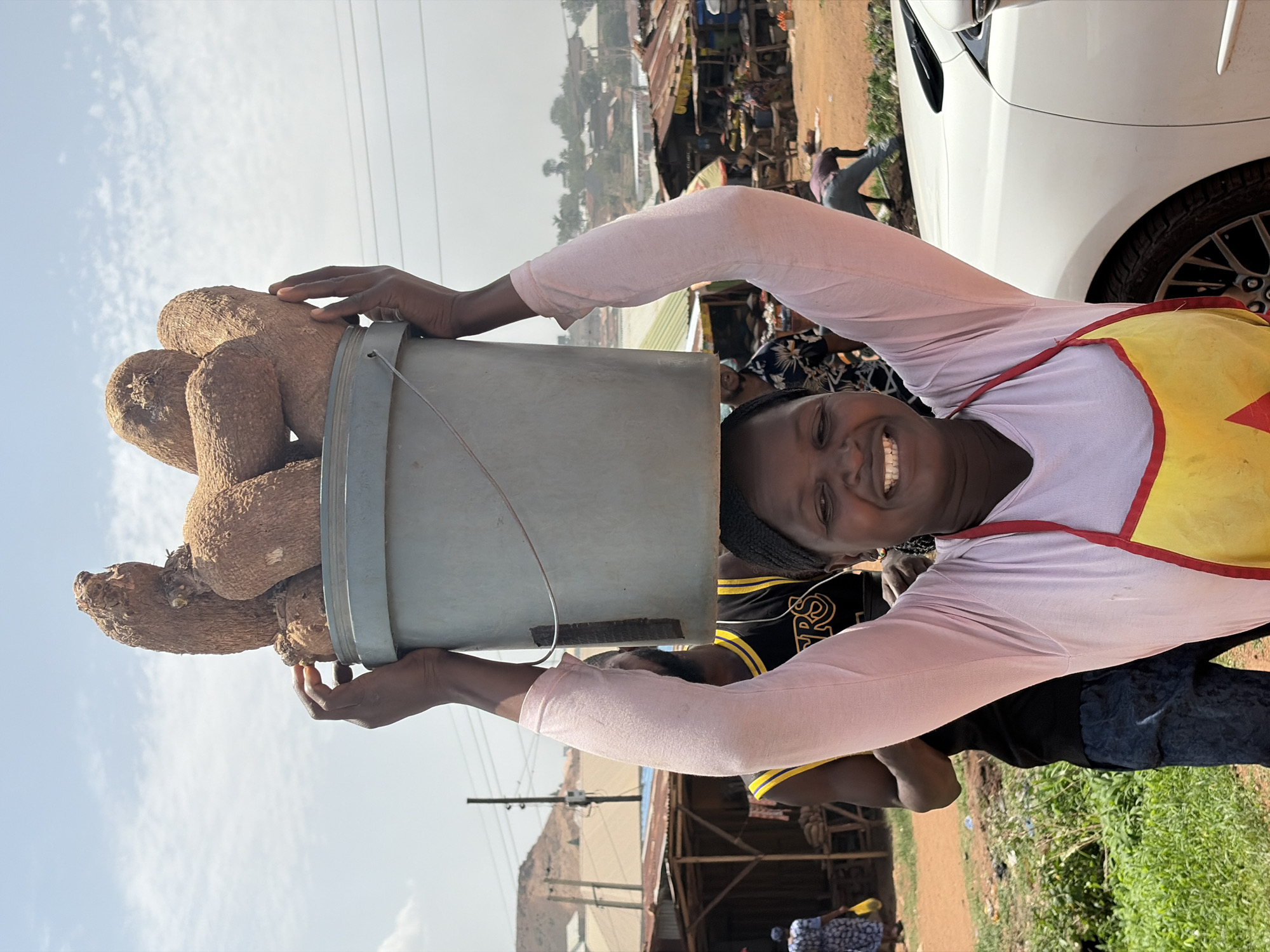 In Barakin Lerawi, Nigeria, a woman smiles directly at the camera, balancing a grey bucket filled with yams on her head. She is wearing a light pink long-sleeved shirt, a yellow apron with red trim, and dark pants. Her arms are raised to steady the bucket.
Behind her, a bustling outdoor market scene unfolds with stalls and people. A white car is partially visible on the right edge of the frame. The background shows buildings, a dirt road, and a blue sky. The environment suggests a typical day in a West African urban setting.