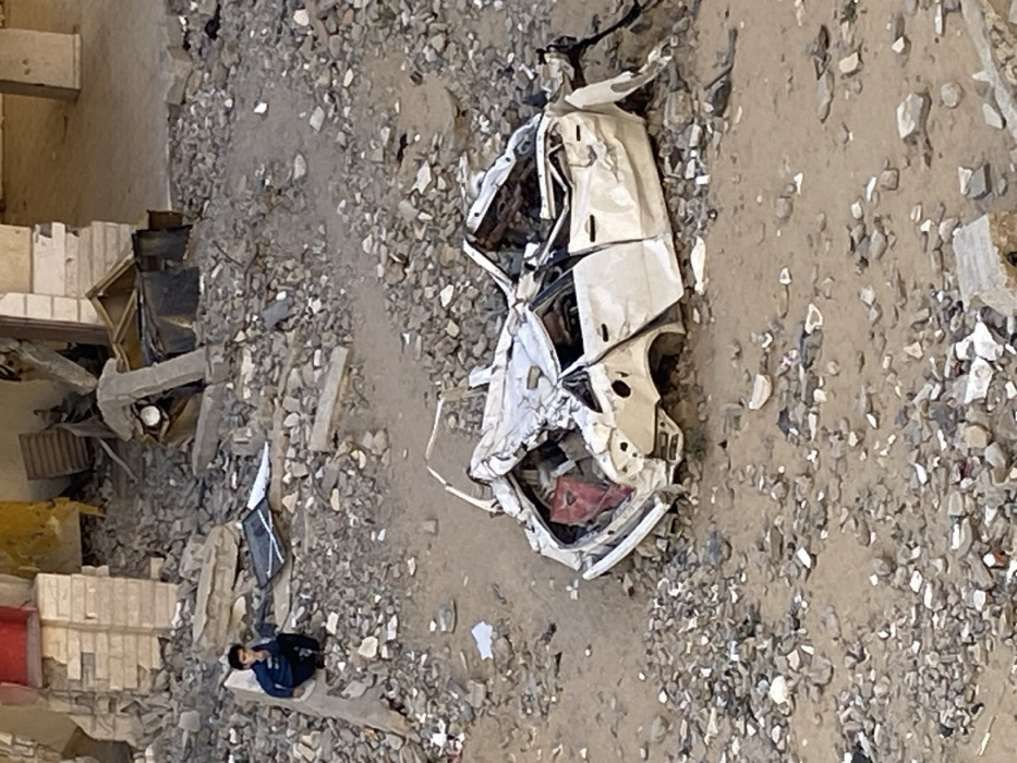 The image captures an outdoor scene of significant destruction and debris. The ground throughout the frame is covered in a mixture of dirt, dust, and various sizes of rubble, including rocks, shattered concrete, and fragmented building materials.
A prominent feature in the middle-right of the image is a heavily damaged white vehicle. The car is severely crushed and mangled, its original shape barely discernible. Its body panels are torn and twisted, the roof appears caved in, and the interior is exposed, revealing warped metal and some red material, possibly upholstery or an item inside. The vehicle lies partially on its side or overturned amidst the extensive debris, indicating a catastrophic impact or collapse.
In the bottom-left section of the image, a young male figure is visible, sitting on a low, light-colored piece of debris or a concrete block. He is dressed in a dark blue long-sleeved shirt and dark trousers. His posture suggests he is looking downwards or slightly ahead, and his expression or demeanor is not clearly visible but implies contemplation or weariness. A dark object, possibly a backpack or a bag, is situated on the ground next to him.
On the far-left side of the image, there are more remnants of what appear to be demolished or destroyed structures, including stacks of bricks or concrete blocks and fragmented wooden or metal elements, further contributing to the scene of ruin. The lighting suggests natural daylight, providing a clear view of the devastation. There is no visible text in the image.
