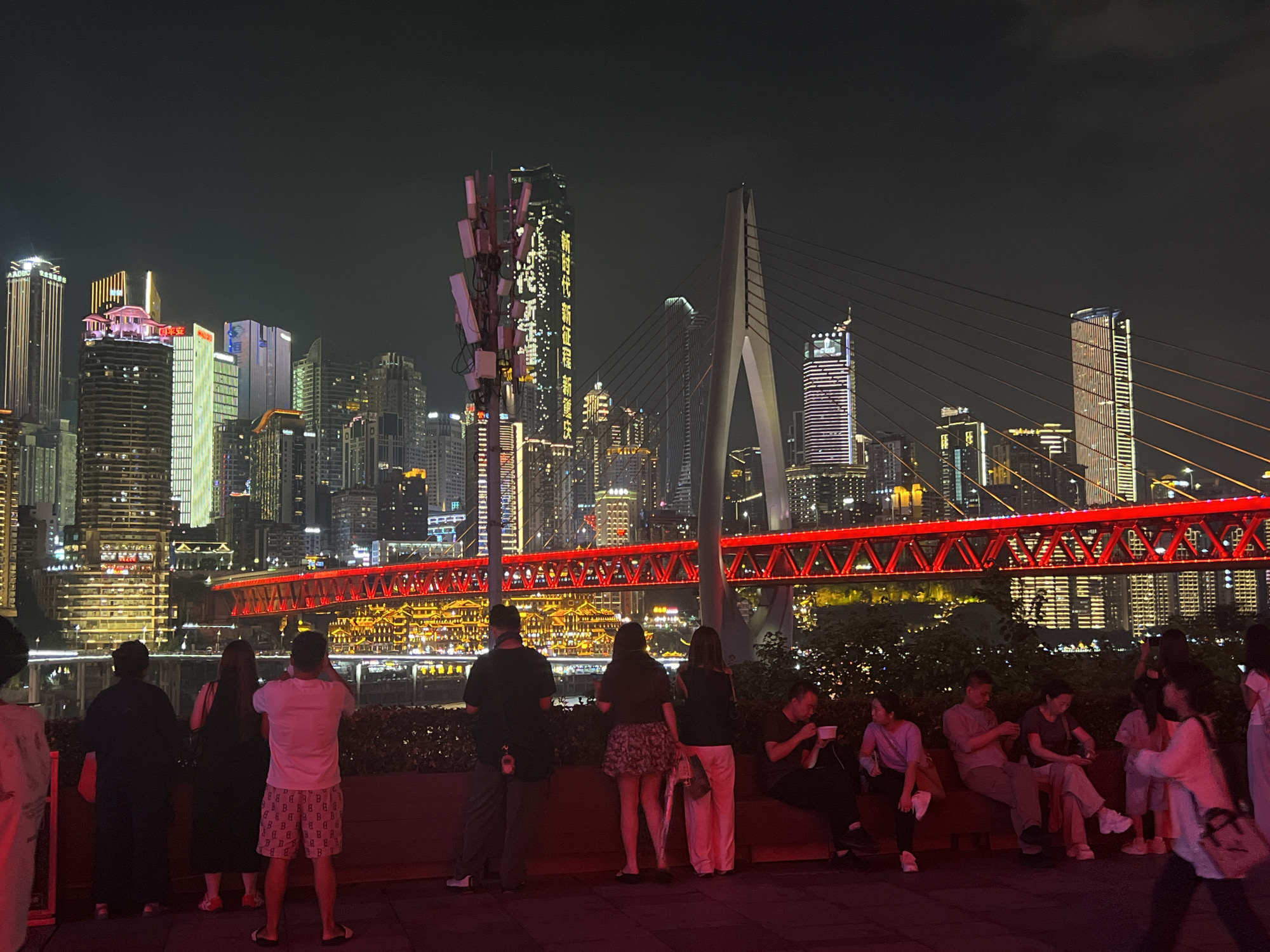 The image depicts a nighttime scene in Jiangbeicheng Street, China, with a prominent, illuminated red bridge spanning across a body of water. A modern cityscape with numerous tall buildings, many of which are lit up with various colors, serves as the backdrop. In the foreground, a group of people are gathered, some standing and observing the view, while others are seated on benches. Several individuals are captured in candid moments, such as taking photographs with their phones, talking, or simply resting. The atmosphere appears to be relaxed, with the artificial lights of the city and bridge creating a vibrant and picturesque ambiance. A telecommunications tower with multiple antennas is also visible on the left side of the frame. The ground in the foreground is a dark, textured surface, possibly a walkway or plaza.