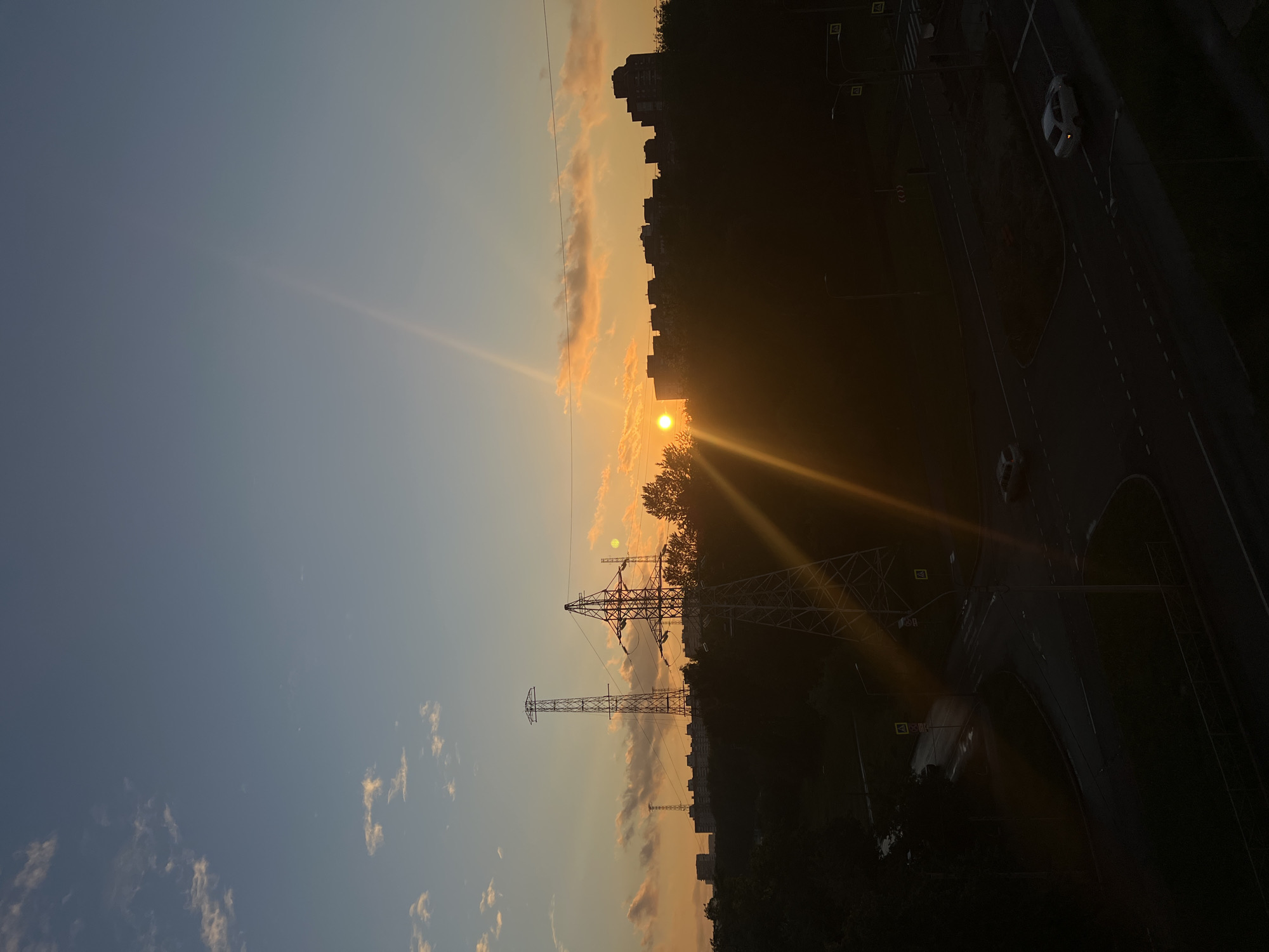 The image captures a sunset over a city, likely Saint Petersburg, Russia, given the context. The sun, a bright orb, is setting on the horizon, casting a warm, orange glow across the sky and illuminating the clouds with golden hues. A distinct sunbeam pierces the sky at an angle. In the foreground and midground, silhouettes of trees and buildings are visible against the vibrant sky. Power lines and tall transmission towers stand out as stark structures against the natural backdrop. The lower part of the image shows a dark, urban environment, possibly a road intersection with streetlights and cars. The scene suggests a typical urban evening, with the natural beauty of the sunset juxtaposed with the man-made elements of the city.