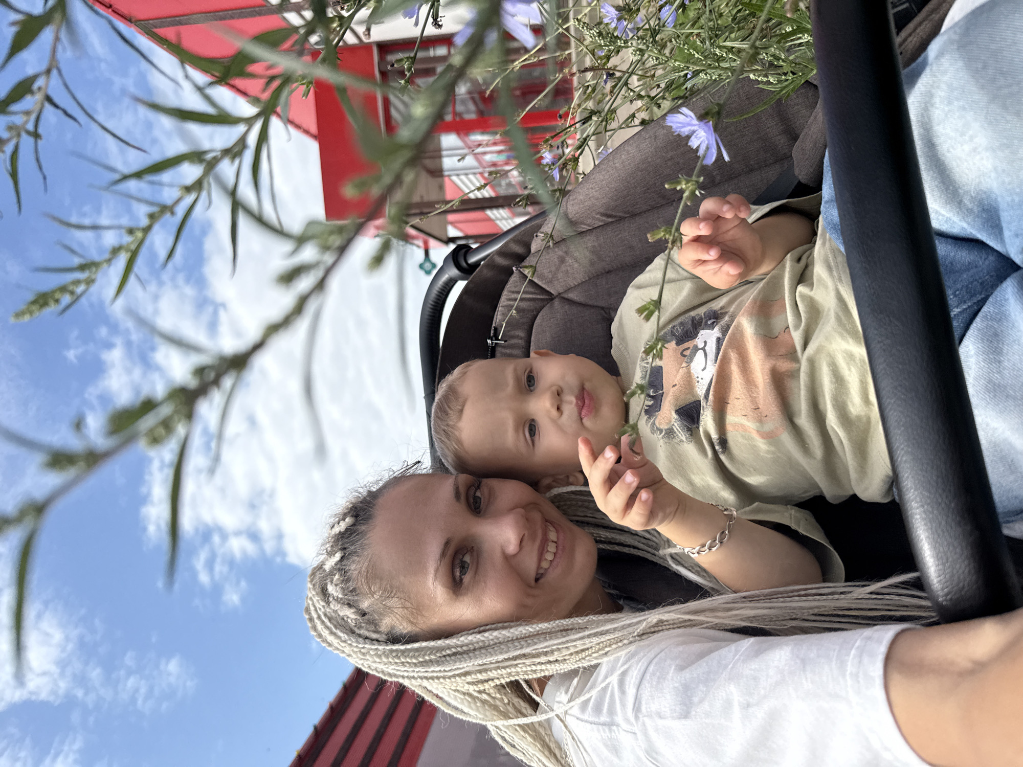In Rusanovo, Russia, a woman with blonde braided hair wearing a white t-shirt is taking a selfie with a young child in a stroller. The child, wearing a light green shirt with a lion graphic, is sitting in the stroller. They are positioned in front of a red building with white trim. Lush green plants and blue flowers are in the foreground, partially obscuring the view. The sky above is blue with scattered white clouds. The image captures a casual outdoor moment.