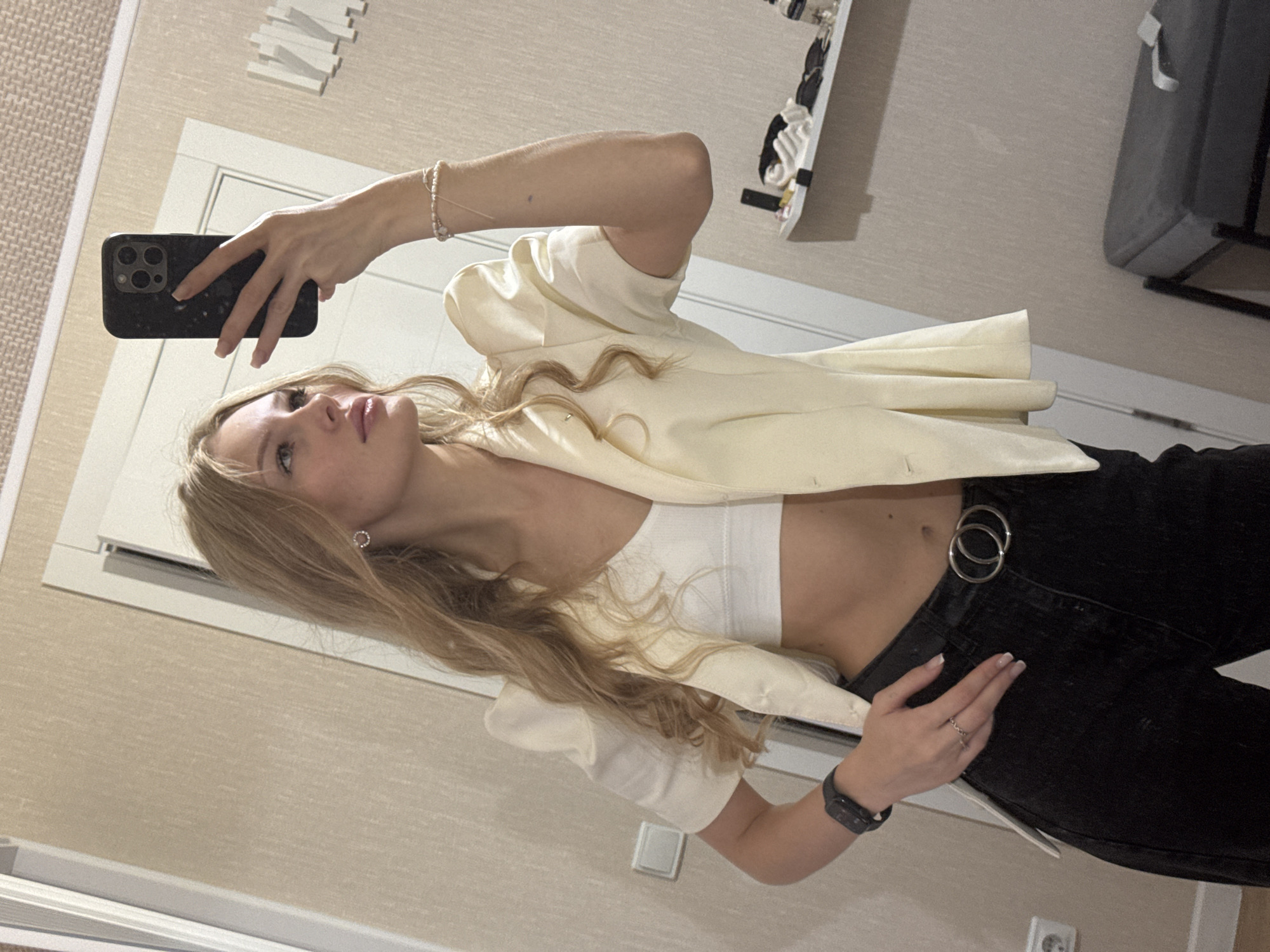A young woman with blonde hair is taking a mirror selfie indoors. She is wearing a white crop top, a light yellow blazer with puffed short sleeves, and black jeans with a double-ring belt. She has on a dark smartwatch and a ring on her left hand, and a beaded bracelet on her right wrist. Her right hand is holding a black smartphone. The background appears to be a room with textured beige walls, a white door, and a shelf with various items. There is no indication of the city of Perm, Russia, in the image.