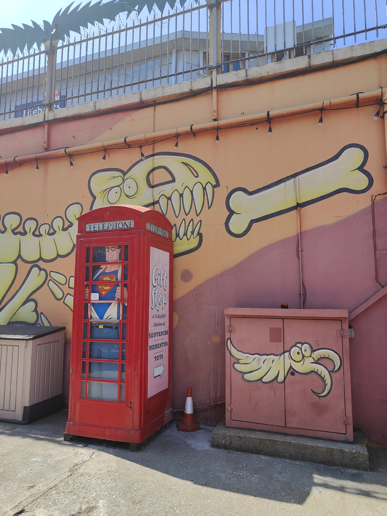 This image shows a red telephone box with a figure dressed as Superman inside, located in Southend-on-Sea, United Kingdom. The telephone box is positioned in front of a mural painted on a wall. The mural depicts a large, cartoonish yellow dinosaur skeleton and a yellow bone. To the right of the telephone box is a pink metal cabinet with a cartoon drawing of a yellow creature with large eyes and tentacles on its doors. A traffic cone is visible on the ground in front of the telephone box. A light tan storage unit is visible to the left of the telephone box. The scene is outdoors, with a concrete ground and a metal fence with spikes at the top in the background. The sky is clear and blue.