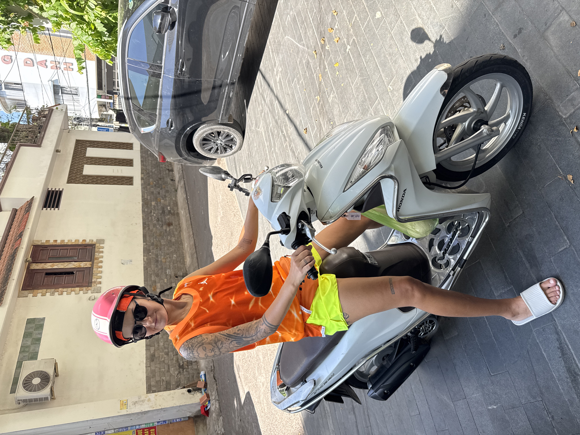 In South Quy Nhon Ward, Vietnam, a woman with tattoos on her arms and legs sits astride a white Honda scooter. She is wearing a pink and white helmet, sunglasses, an orange tank top with a tie-dye pattern, and bright yellow shorts. The scooter is parked on a gray tiled sidewalk, with fallen leaves scattered around. Behind the scooter, a black SUV is visible, and further in the background, there are buildings, including one with a sign that reads "NG DANG H." The scene is illuminated by bright sunlight, casting shadows on the ground.