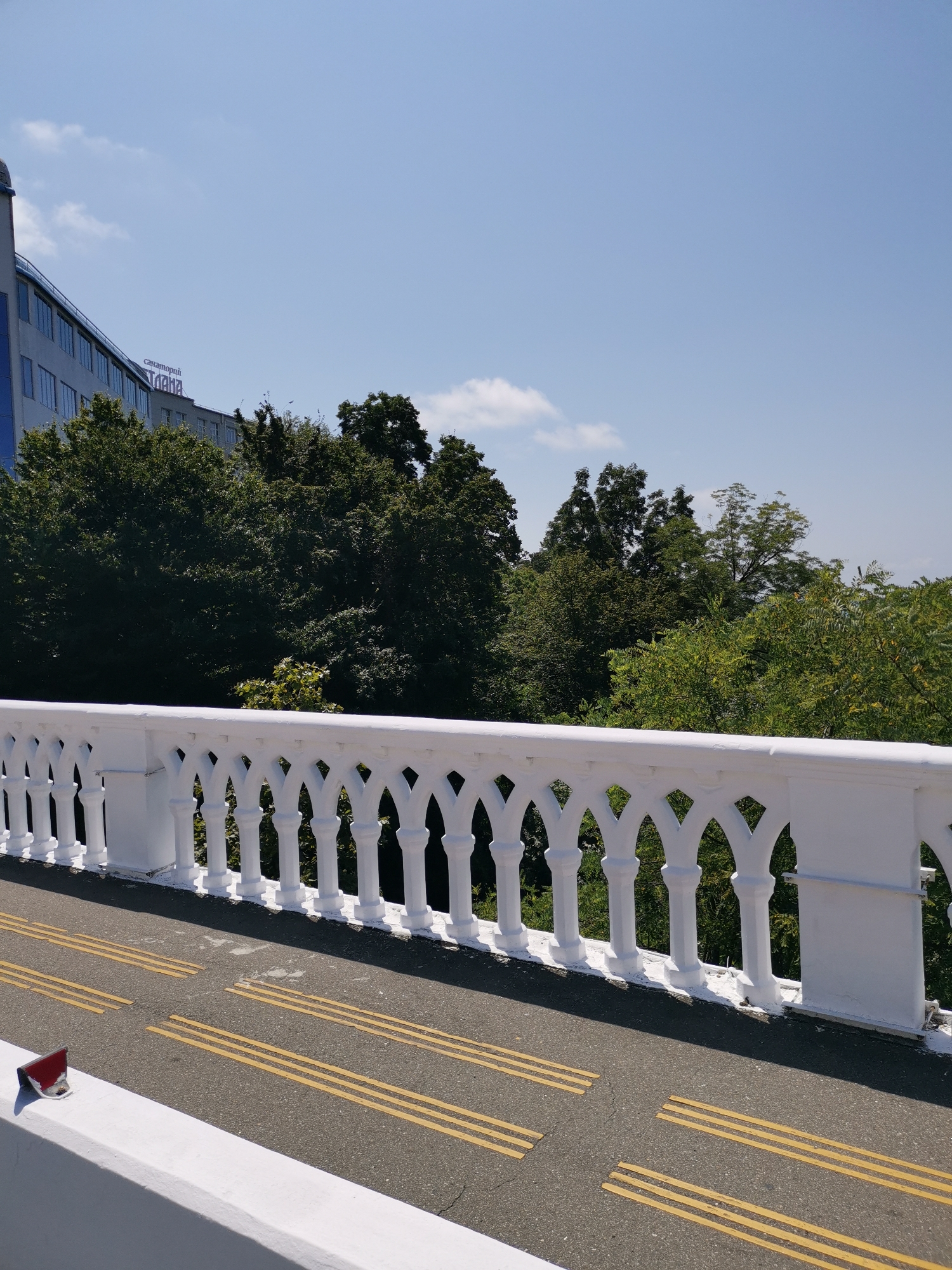 The image shows a view of a white, ornate balustrade on a bridge in Sochi, Russia. The balustrade features decorative arches and columns, painted white. A paved walkway or road runs alongside the balustrade, marked with yellow tactile paving lines. In the background, there is a large, multi-story building with blue-tinted windows, and a sign with Cyrillic lettering that reads "санаторий «Ивушка»" (Sanatorium "Ivuska"). Beyond the building and the balustrade, a dense canopy of green trees is visible under a clear blue sky with a few wispy clouds. The scene appears to be a sunny day.