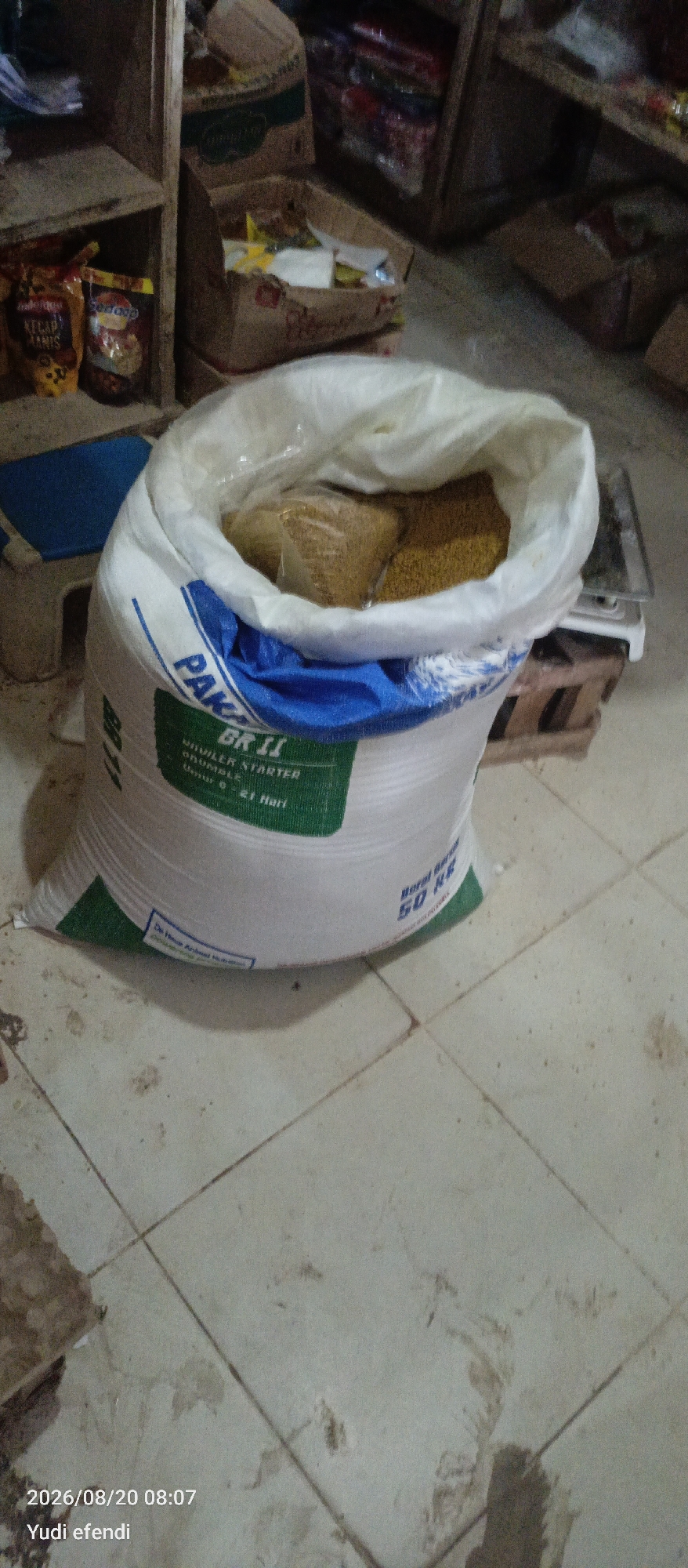 The image depicts the interior of a room, likely a storage area or shop, with a focus on a large, open sack of animal feed. The sack is white with blue and green lettering. The visible text on the sack includes "PAK" followed by "BR II," "UNIVILER STARTER," "GRUMBLE," and "Usia 0 - 21 Hari." Another line of text reads "Berat Bersih 50 KG." The sack appears to be partially filled with a granular, yellowish substance.
In the background, shelves are visible, stocked with various items, including what appear to be food products in pouches and boxes. Two such pouches are on the left, with labels partially visible, one reading "KECAP MANIS." Other boxes are stacked, some appearing damaged. A blue plastic object is partially visible under the sack. A white electronic scale is situated behind the sack, resting on wooden supports. The floor is tiled in a grid pattern and appears somewhat dirty.
The overall scene suggests a commercial or agricultural setting where animal feed is stored or sold. The presence of the scale and the packaging of the feed indicate a business operation. The lighting is dim, contributing to a somewhat grainy appearance of the image. The timestamp at the bottom of the image indicates it was taken on August 20, 2026, at 08:07, and the name "Yudi efendi" is associated with it, possibly the photographer or the owner. There is no direct indication of Rawajitu Selatan, Indonesia within the visual information of the image itself.