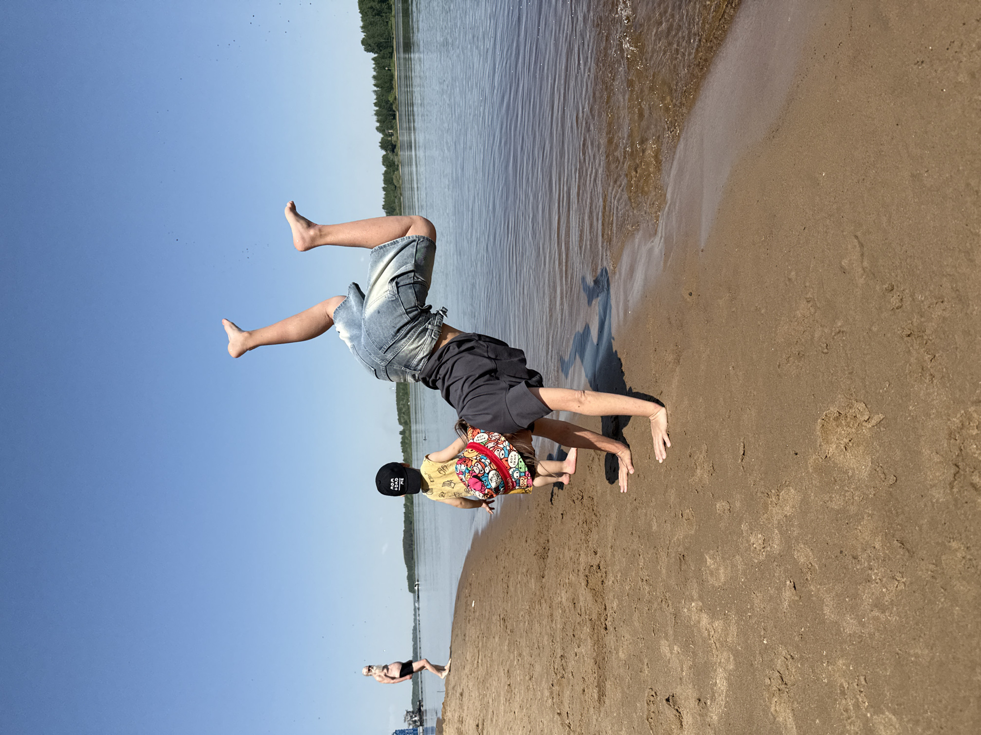 The image shows two children and an adult on a beach, near water, in Rusanovo, Russia. One child is upside down and in mid-handstand, wearing denim shorts and a dark t-shirt. The other child is standing upright, wearing a yellow patterned shirt, a black baseball cap, and a colorful backpack. An adult figure, wearing dark swimming attire, stands further down the beach toward a distant building. The beach consists of sand, and the water is in the background. The sky is light blue.