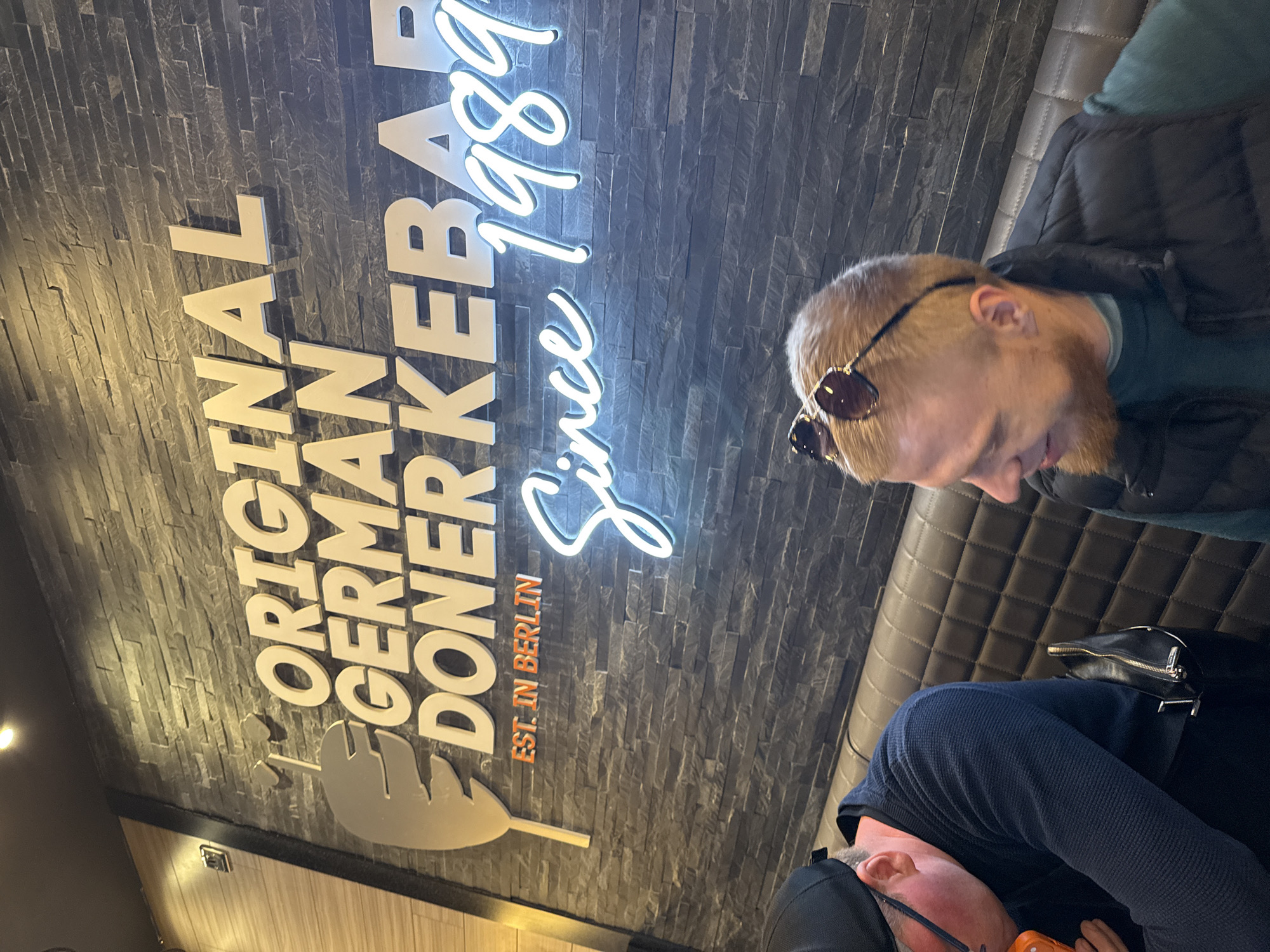 Two people are sitting inside a restaurant. One person is wearing sunglasses and a black vest, while the other is wearing a dark blue sweater. Behind them is a brick wall with a sign that reads "Original German Doner Kebab. Est. in Berlin. Since 1989." The restaurant is in Great Yarmouth, United Kingdom.