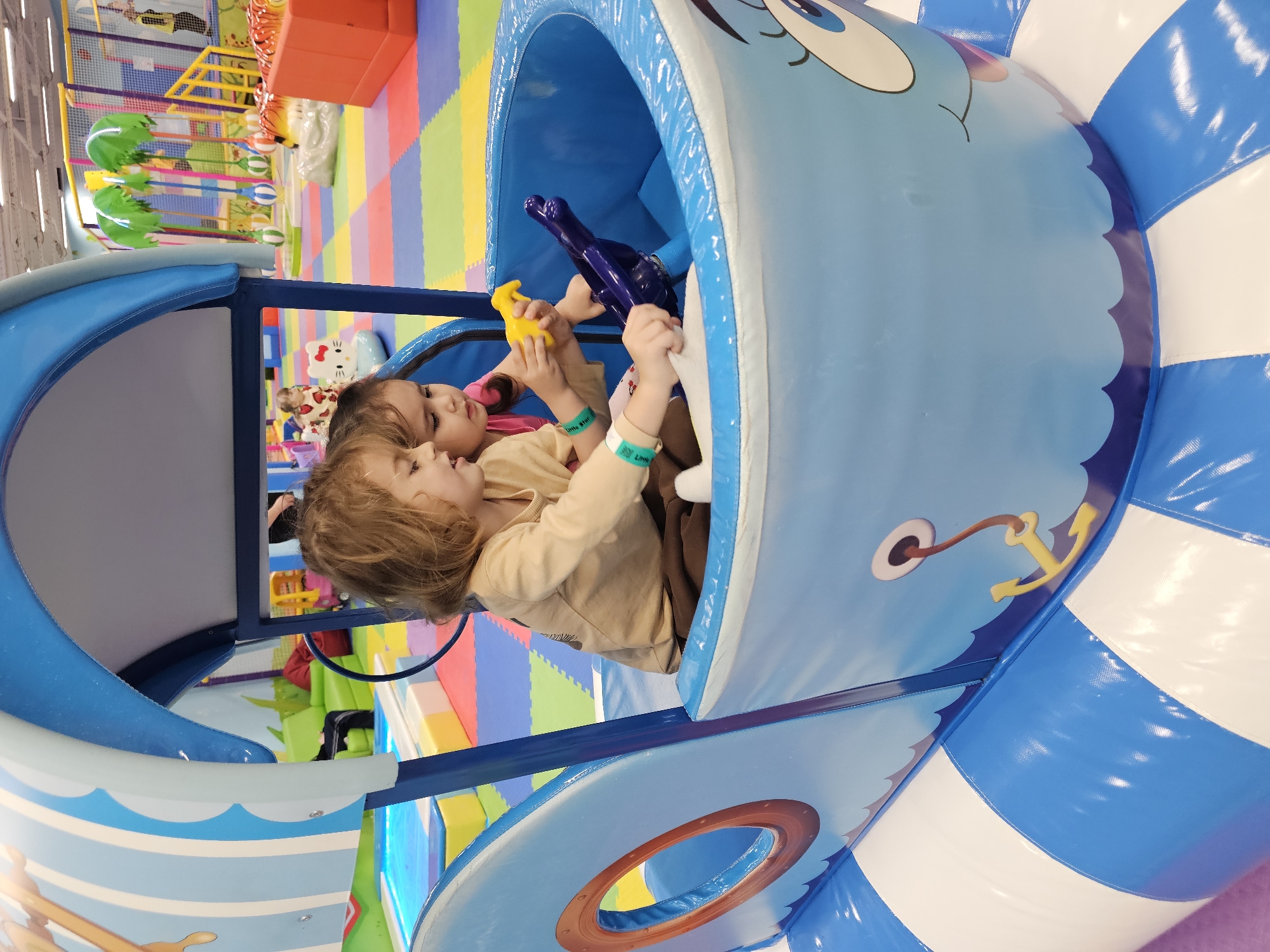 Two young children are inside a blue, inflatable boat-shaped play structure. The child closest to the viewer is wearing a beige long-sleeved shirt and brown pants. The other child is wearing a pink shirt. Both children have wristbands on their left arms. The children are holding a purple steering wheel and a yellow object. The background shows colorful padded flooring and other play structures. The location is likely an indoor playground in Khimki, Russia, based on the metadata.